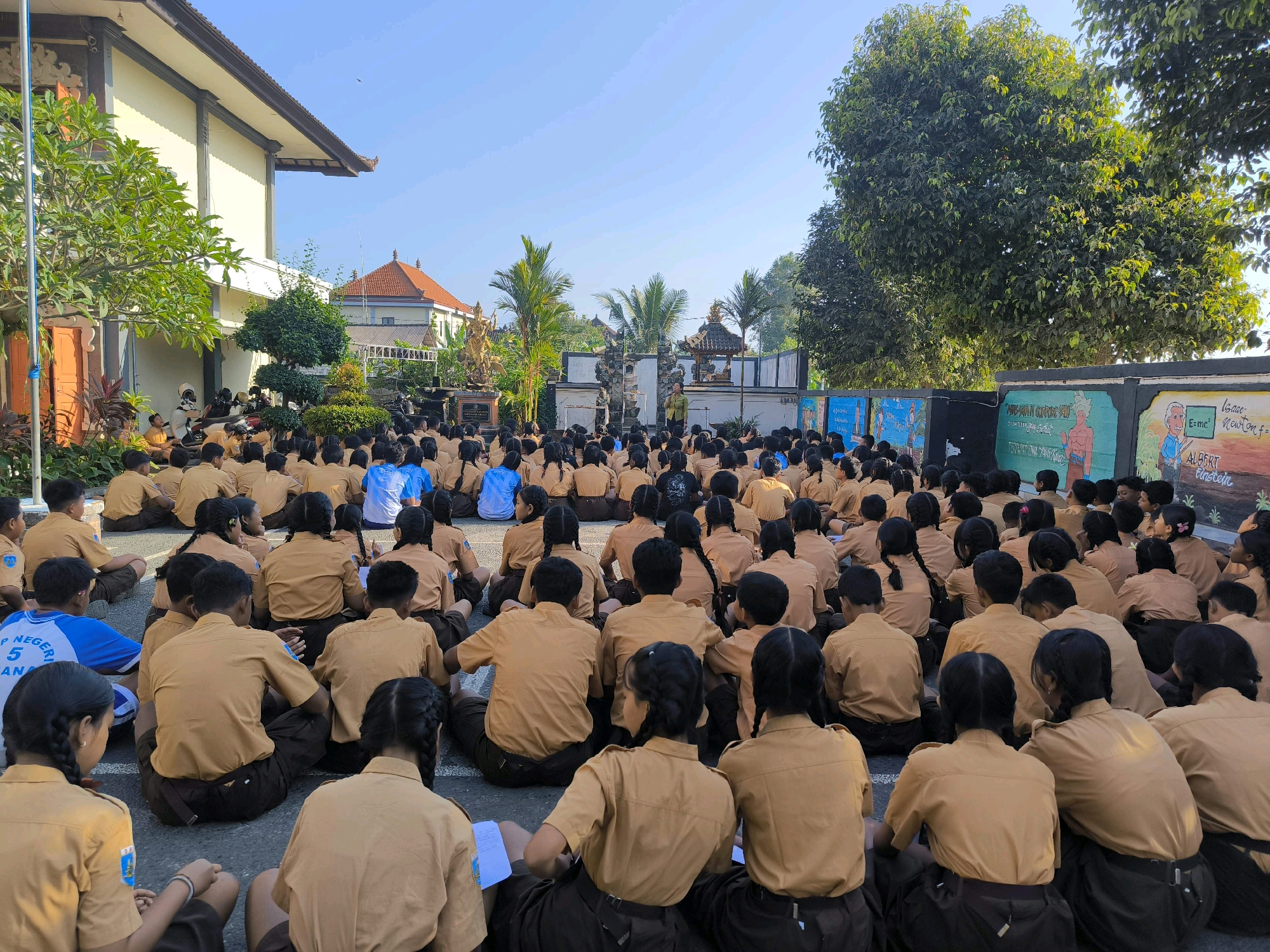 In Tabanan, Indonesia, a large group of schoolchildren in tan uniforms are sitting on the ground facing a stage. The children, with diverse hair colors and textures, are attentive and engaged, with some holding papers. The stage area features a traditional Balinese gate, statues, and murals, suggesting a cultural or educational event. Behind the stage, palm trees and lush greenery contribute to the tropical atmosphere. The background includes a building with a red-tiled roof and a clear blue sky, indicating it is a sunny day. The overall scene depicts a community gathering, likely at a school or public space, focused on an outdoor presentation.