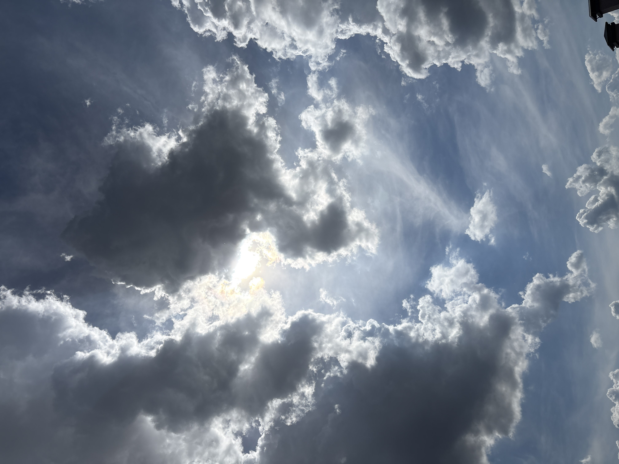 This is a vertical, full-frame, eye-level shot of a cloudy sky above Sofia, Bulgaria. The sun is partially obscured by dark, dramatic clouds, creating a sense of depth and contrast. The clouds are varied in texture and density, with some appearing wispy and thin, while others are thick and ominous. The sky transitions from a bright, sunlit area in the upper center of the frame to a deeper blue towards the edges. No people or man-made structures are visible, except for the very top of two chimney structures in the bottom right corner. The image captures a moment of dynamic atmospheric conditions.