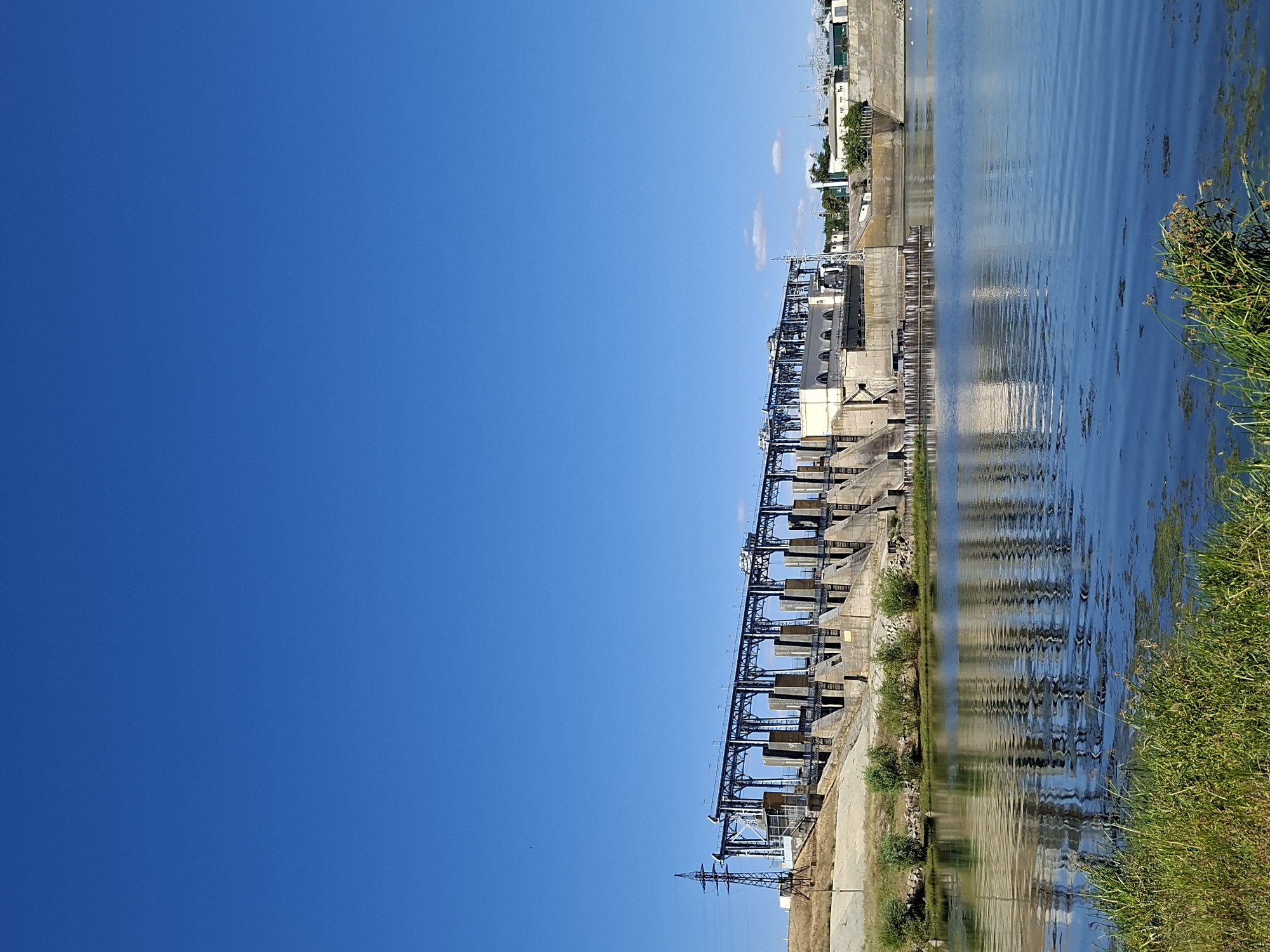 The image shows a large hydroelectric dam on a wide, calm river under a clear blue sky. The dam, constructed of concrete and steel, features multiple spillway gates supported by a steel superstructure with a walkway. A tall, lattice-style electrical pylon stands to the left of the dam. The river reflects the dam's structure and the sky. The foreground of the image shows green grass and some aquatic vegetation along the riverbank. In the background, beyond the dam, a few buildings and some trees are visible, suggesting an urban or industrial area. The overall scene depicts a functional piece of infrastructure in a natural setting. This scene is located at the hydroelectric power plant in the city of Ustia, Moldova. There are no people visible in the image.