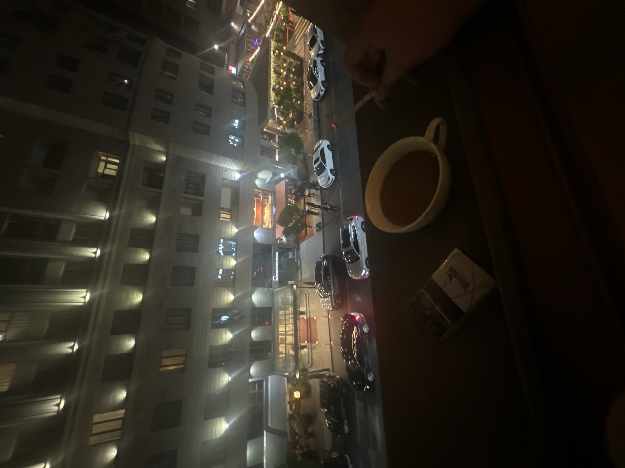 The image appears to be taken at night from a high vantage point in Yerevan, Armenia. The view looks down onto a street with several cars parked along the side. There is a tall building with many windows visible. A person is holding a lit cigarette, while a mug of liquid and a pack of cigarettes are on a table nearby.