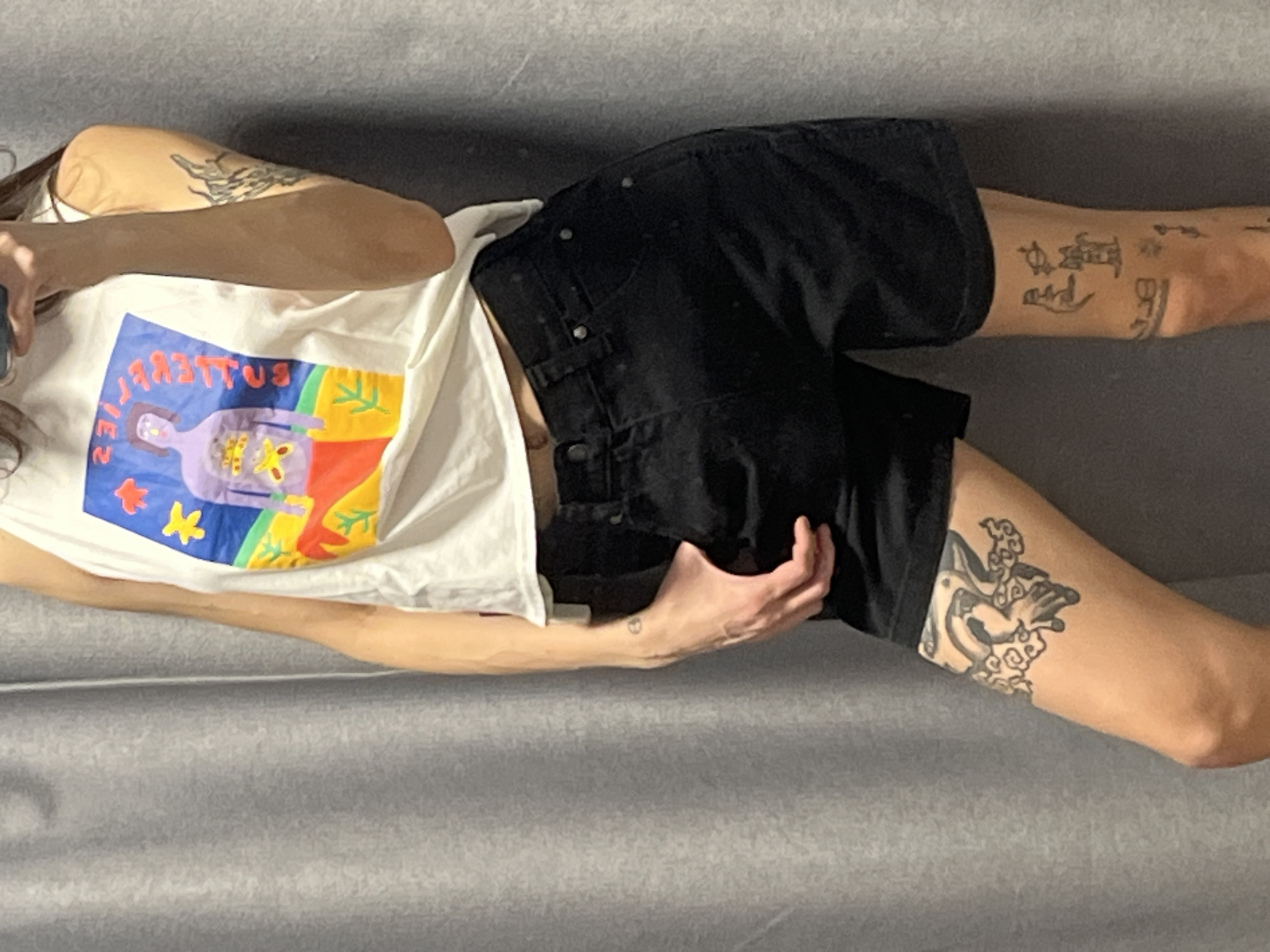 An individual, appearing female, is captured in a waist-down, mirror-style photograph within an indoor setting. The subject is wearing a white sleeveless tank top featuring a colorful graphic print depicting an inverted purple figure with exposed internal organs, standing on a landscape of red, yellow, green, and blue. Inverted Cyrillic text is visible on the shirt. Black denim shorts with a button fly and metal rivets are worn on the lower body. The subject's right hand holds a smartphone, which is used to take the photograph and partially obscures the upper face and head. The left hand is partially inserted into the left pocket of the shorts. Visible tattoos include an abstract dark design on the right bicep/tricep, a prominent dark tattoo of a creature (possibly a frog or toad) surrounded by cloud-like motifs on the left upper thigh, a small dark circular tattoo on the left forearm, and several small, distinct outline tattoos (including a feline figure and a spherical object with a ring) on the right shin/calf. The background consists of a light grey, vertically ribbed fabric curtain on the left, and a textured, lighter grey wall on the right. This environment suggests a retail clothing store fitting room or a private domestic space. The precise location within Krasnoyarsk, Russia, is not identifiable from the image.