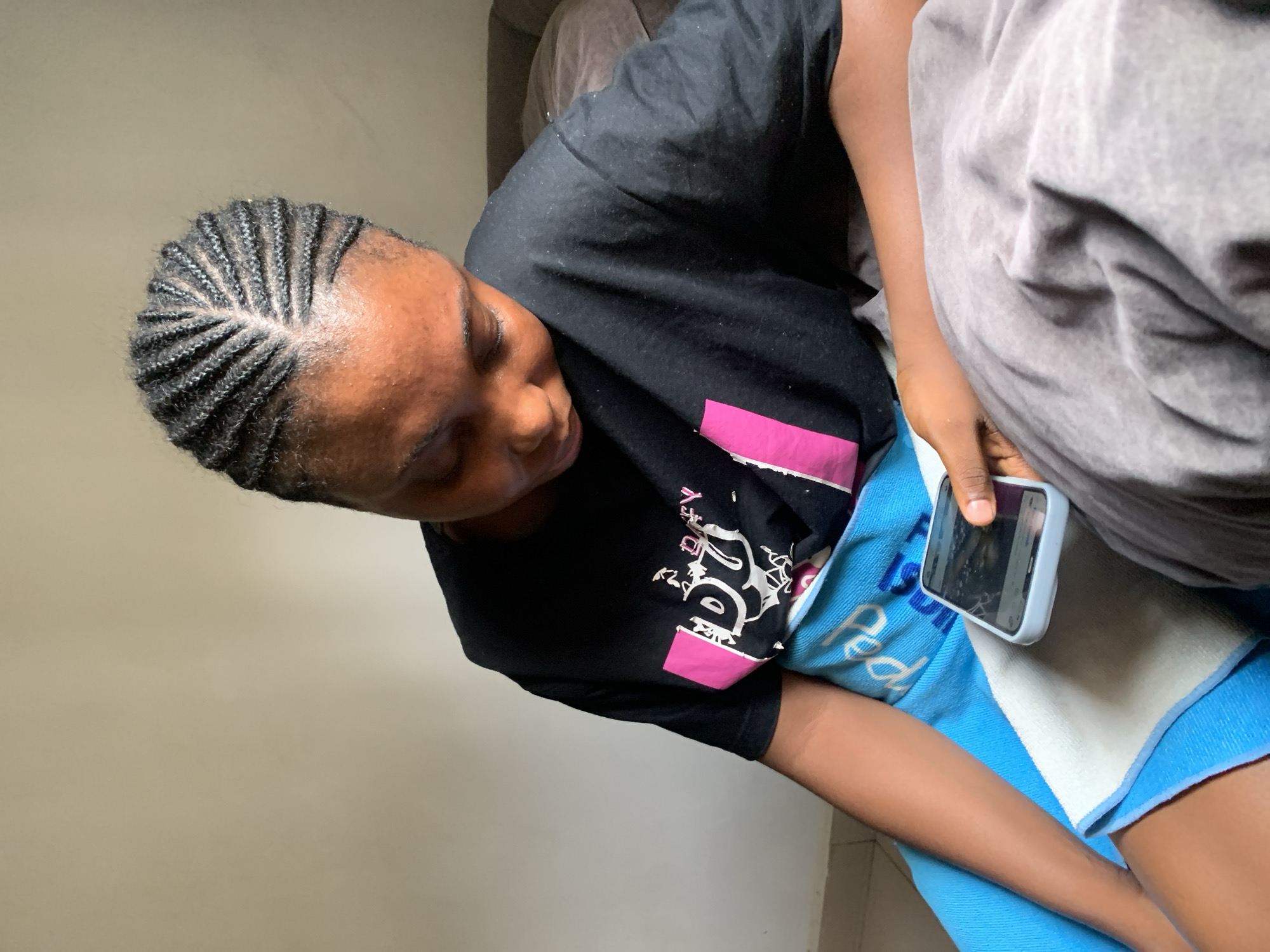 A woman with braided hair sits indoors in Abuja, Nigeria. She is wearing a black T-shirt with a white and pink graphic design. She holds a smartphone in her right hand. A blue and white towel covers her lap. Another individual, partially visible, sits beside her under a gray blanket. The background is a plain, light-colored wall.