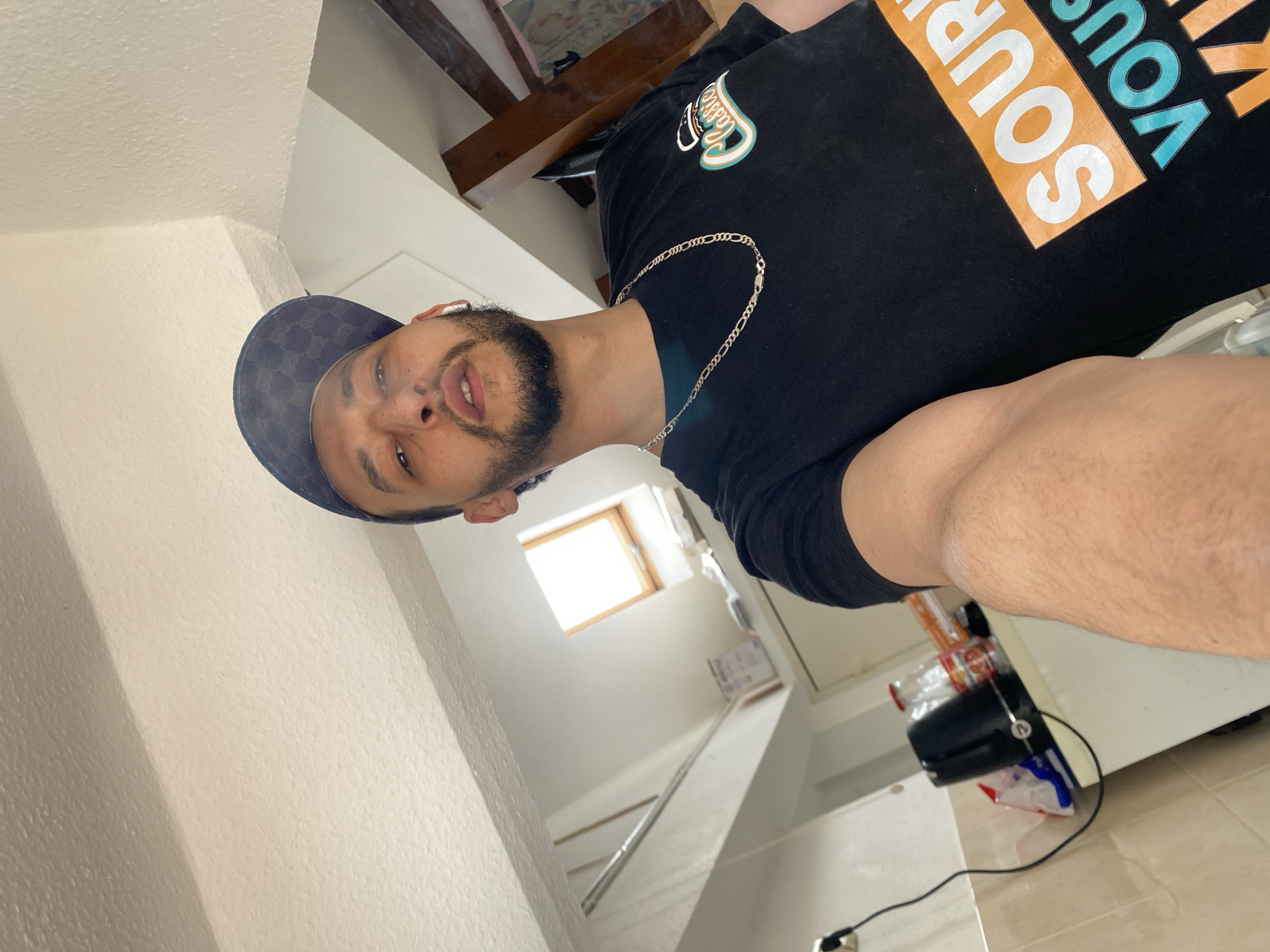 A man wearing a black t-shirt and a patterned baseball cap is taking a selfie indoors. He has a beard and is wearing a silver chain. The background includes a staircase and a window letting in natural light. A vacuum cleaner and other items are visible on the floor. The location is Bischheim, France.