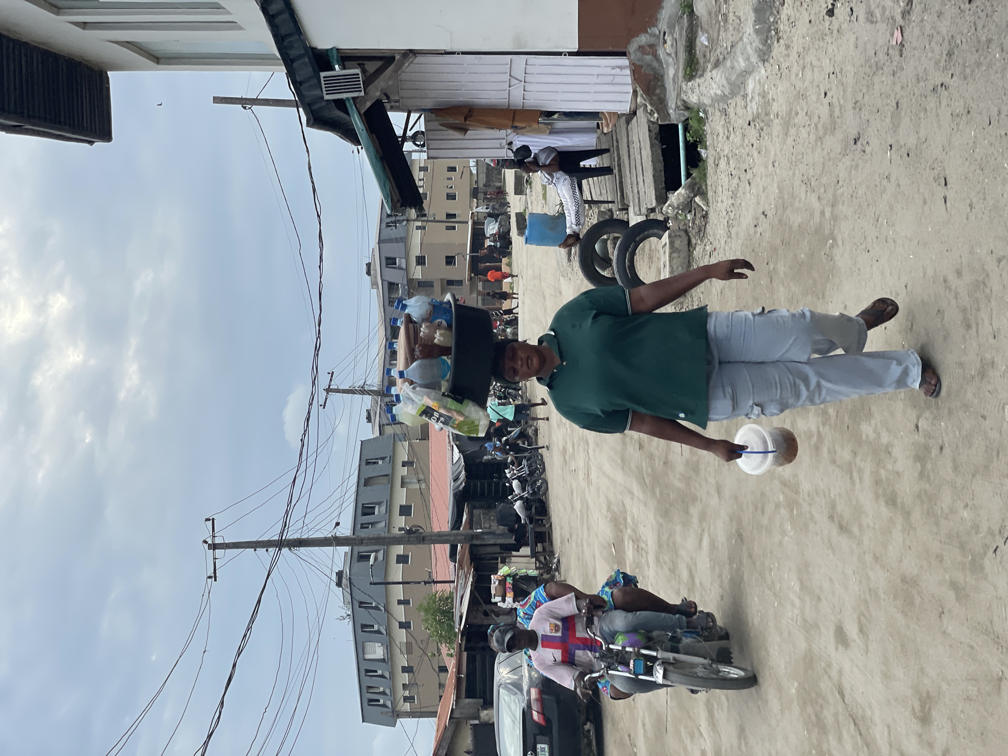 A street scene in Lekki, Nigeria, shows a man balancing a bucket full of plastic bottles and containers on his head, and holding a covered plastic container in his left hand. He wears a green polo shirt and light gray pants. A motorcycle with two riders passes by, one wearing a Barcelona football jersey. Buildings and overhead power lines are visible in the background. The street appears unpaved. In the background there is what appears to be a tire and people outside of a store or gathering space.