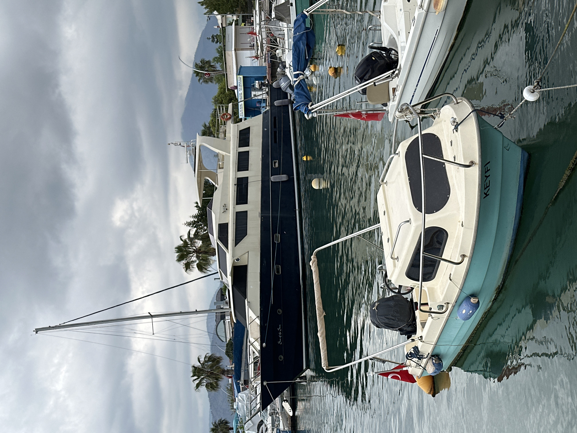 The image depicts a marina scene in Fethiye, Turkey, showcasing a variety of boats docked alongside each other. The boats range from smaller sailboats to larger yachts, all bobbing gently in the water. The water is calm and reflects the cloudy sky above.
Foreground: A small, light-colored boat labeled "KEYFI" is tied to a larger white yacht. A Turkish flag is visible at the stern of the smaller boat. A black bag is on the stern of the boat.
Midground: A large, dark-hulled yacht with white superstructure is docked parallel to the smaller boats. The name "Sea of the Seas" is visible on the hull. The yacht has multiple decks and a large mast.
Background: Buildings and trees line the waterfront. Mountains are visible in the distance, partially obscured by clouds. The sky is overcast, suggesting potentially inclement weather.
Environment: The marina appears well-maintained, with clean water and organized docking arrangements. The overall atmosphere is peaceful and serene.