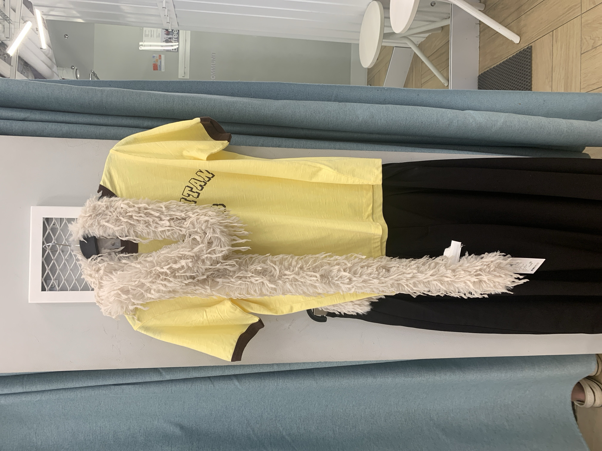 The image shows a clothing display in what appears to be a fitting room or retail space. A bright yellow t-shirt with dark brown trim and a fuzzy, cream-colored scarf is hanging on a rack. Below the t-shirt, a black skirt is visible. The background consists of teal-colored curtains and a white wall. In the background, a reflection shows a mirror, two white stools, and a grey doormat on a light wood floor. The location is identified as Hrodna, Belarus.