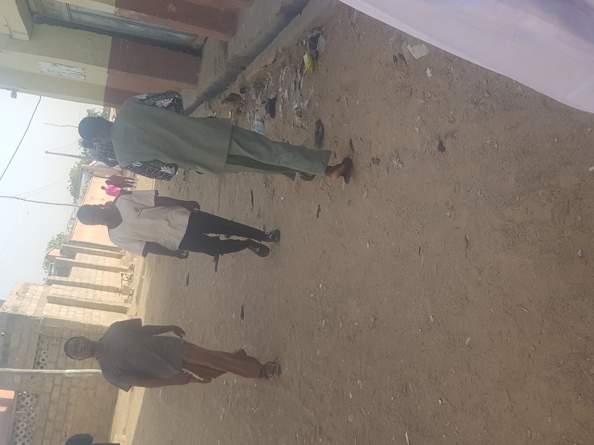 The scene captures an outdoor street view in Dabawa, Nigeria. The ground is mostly dirt with visible debris. Three individuals are prominent in the frame. One is dressed in a green outfit. Another wears a white top with dark pants. The third is in a grayish top and reddish pants. Buildings are visible in the background.