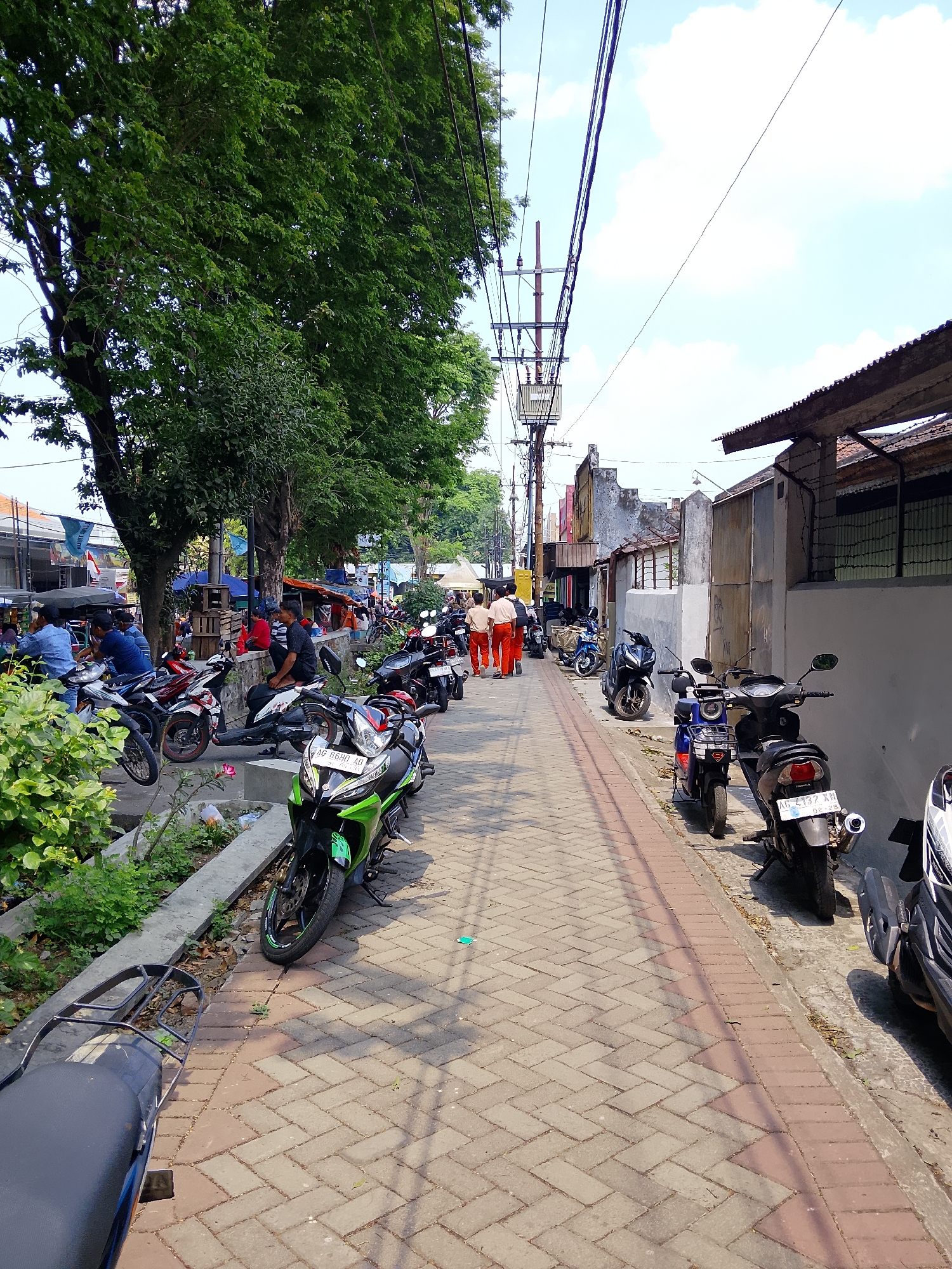 The image depicts a street scene in Kertosono, Indonesia, on a sunny day. A paved walkway runs down the center of the frame, lined with parked motorcycles on both sides. Lush green trees with dense foliage are on the left, casting shadows on the walkway. In the background, several buildings with tiled roofs and grey walls are visible. Electrical poles and wires crisscross the sky, typical of urban infrastructure. A few individuals are present, including two people walking away from the camera in orange pants and white shirts, and others are seen sitting or standing near parked motorcycles and market stalls on the left side of the street. The environment appears to be a commercial or residential area with a lively atmosphere, suggested by the presence of market stalls and multiple motorcycles. The ground is covered in interlocking brick pavers, with some areas of concrete and a curb separating the walkway from the vegetated area on the left.