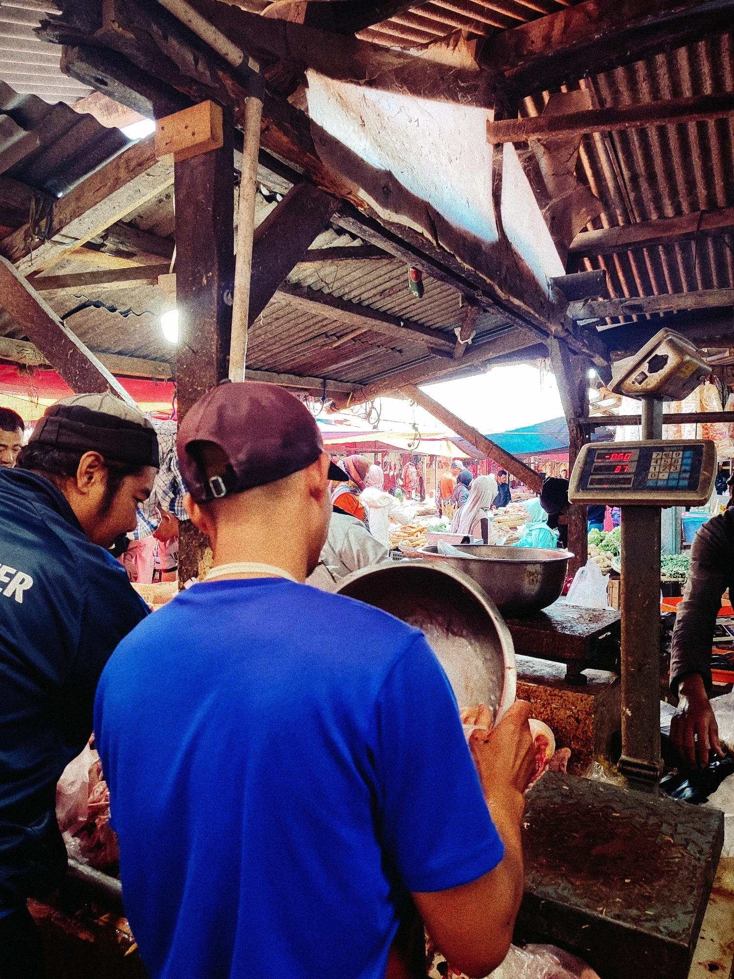 The image depicts a busy marketplace scene, likely in the city of Garut, Indonesia, with an emphasis on a butcher's stall. In the foreground, a man wearing a blue t-shirt and a maroon baseball cap is seen from behind, holding a large metal bowl and appearing to be in the process of preparing meat. To his left, another man in a blue jacket with white lettering that partially reads "ER" is also partially visible.
The environment is characterized by a rustic, open-air structure with a corrugated metal roof supported by wooden beams. Sunlight filters through gaps, illuminating the scene. In the background, other vendors and customers are present, with stalls displaying produce and goods. A prominent feature is a vintage weighing scale with a digital display showing "-0.60" and "31". Several large metal basins and plastic bags containing what appears to be raw meat are on display and in use. The overall impression is one of a functional and active marketplace where transactions and food preparation are taking place.