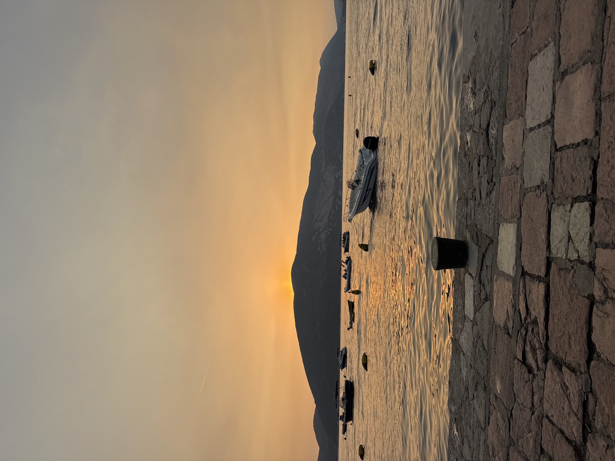 The image depicts a waterfront scene at sunset. In the foreground, a stone-paved walkway leads to the water's edge, with a dark, cylindrical bollard on the right. The water is calm with gentle ripples reflecting the warm orange and yellow hues of the sky. Several boats are scattered across the water, some appearing to be anchored. In the middle ground, a small white motorboat with blue trim is visible. Further in the distance, a chain of dark, silhouetted mountains forms the horizon. The sky is a gradient of soft orange and yellow, with the sun partially obscured by the mountains, casting a warm glow. A faint contrail from an airplane is visible in the upper left quadrant of the sky. The overall atmosphere is serene and tranquil.