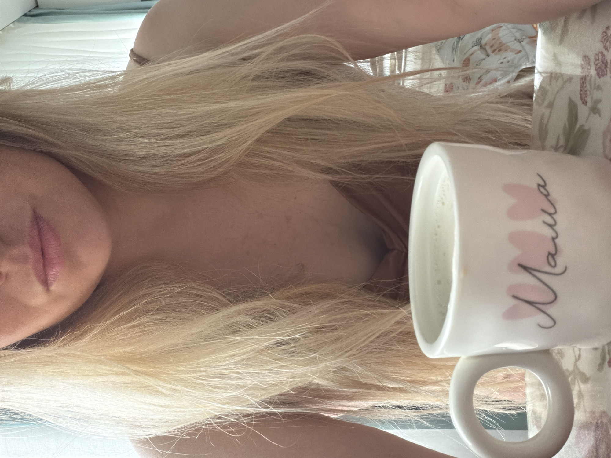 A close-up, low-angle, head-and-shoulders portrait of a fair-skinned, blonde woman with long hair partially obscuring her face and chest. Only her lips and the left side of her face are fully visible. She is wearing a tan or beige garment. A white ceramic mug with the word "Mama" written vertically in black cursive and adorned with pink hearts is positioned in the right of the frame. The mug contains a light-colored liquid. The background appears to be a bed with floral-patterned bedding. The presence of light-colored walls and curtains indicate an indoor setting, such as a bedroom. There is no information to determine if the location is Votkinsk, Russia.