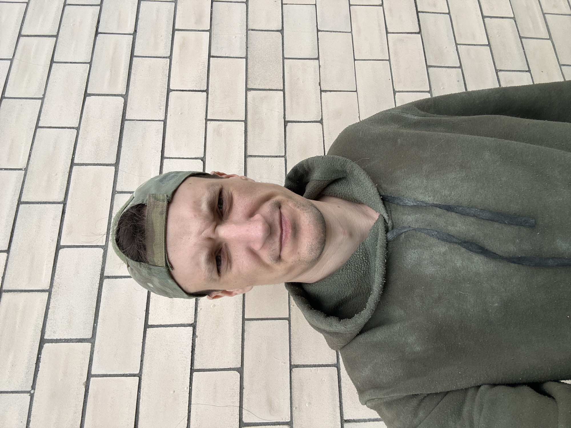 A man wearing a green hooded sweatshirt and a camouflage baseball cap is standing in front of a light-colored brick wall. The man's face is visible, and he has a stern expression. His hair is dark brown, and he has a light beard. The wall is made of rectangular bricks arranged in a staggered pattern, with dark grout lines between them. The man is looking directly at the camera. The overall impression is that of a casual outdoor portrait.