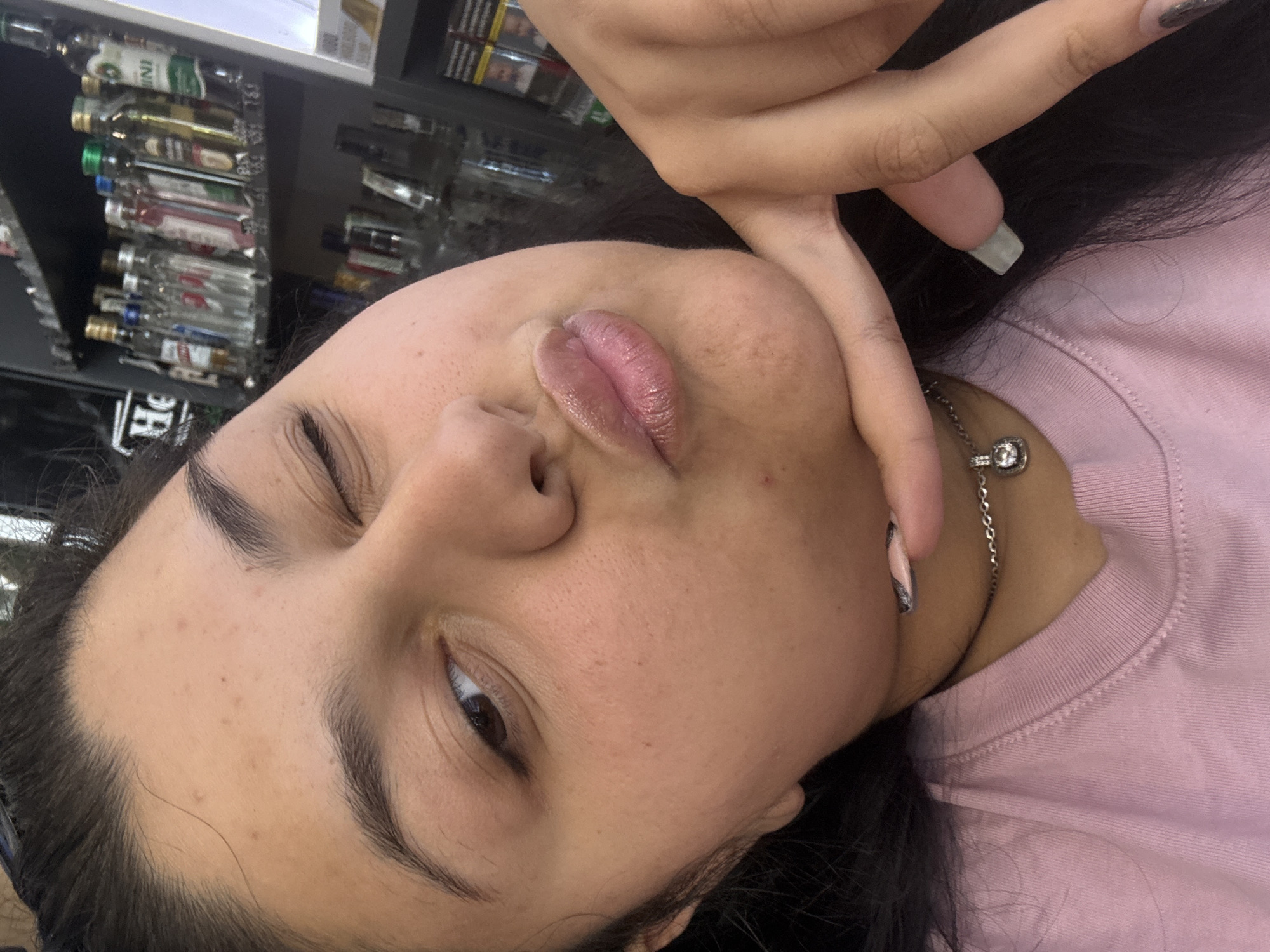The media file is a close-up, head-and-shoulders selfie of a young person. The person is lying down, with their head tilted. Their face is the primary subject, with their lips slightly parted and eyes closed, giving a relaxed or playful expression. A hand with manicured nails is resting near their face, obscuring part of their cheek. A silver necklace with a clear pendant is visible around their neck, resting on a pink t-shirt. The background appears to be a retail setting, possibly a liquor store, with shelves of miniature bottles of alcohol visible. The lighting suggests an indoor environment, potentially during daylight hours. There is no discernible weather or time of day information, and no text is clearly legible in the image. The setting implies a casual moment captured in a commercial space.