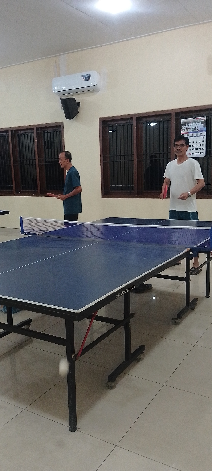 The image depicts an indoor setting, likely a recreation room, where two men are playing table tennis. The room has light-colored walls and a tiled floor. On the wall, an air conditioning unit is visible, along with barred windows. A calendar hangs on the wall above the right-side player. A blue table tennis table with a net is centrally located, with the two men positioned on either side, holding paddles. The man on the left wears a teal t-shirt and black pants, while the man on the right wears a white V-neck t-shirt and dark shorts. The atmosphere appears casual and engaged, suggesting a friendly game. The table itself is marked "DOUBLE FISH". The scene implies a location within Kudus, Indonesia, given the context of the request.