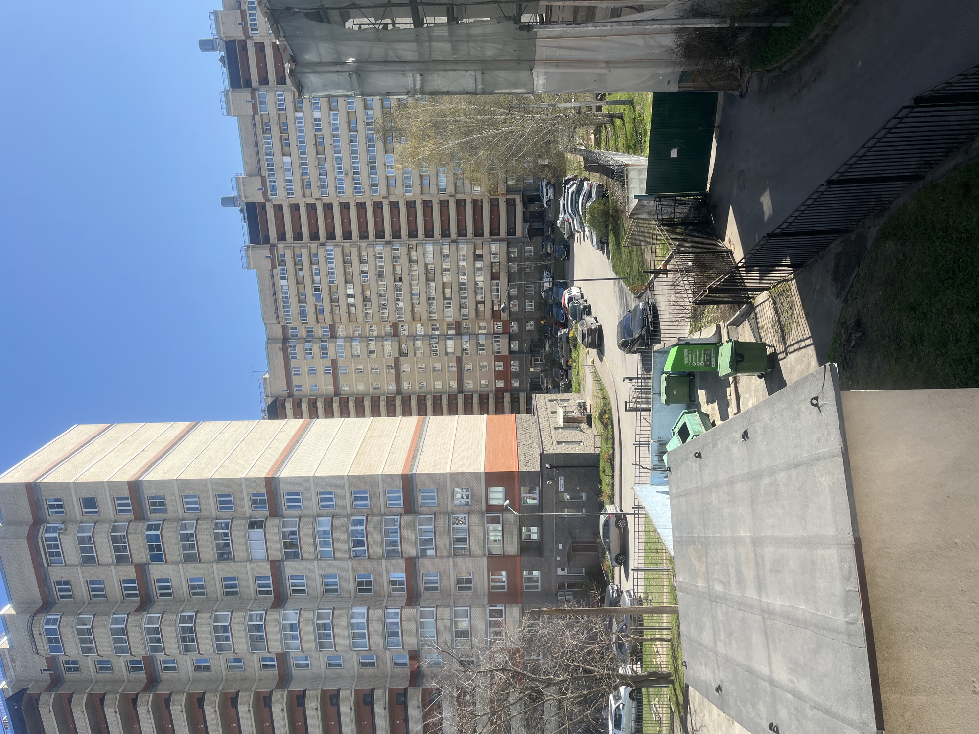 This image presents a bright, sunny daytime view of a residential area in Saint Petersburg, Russia. The scene is dominated by two prominent multi-story apartment buildings under a clear blue sky.
The building on the left is tall, featuring a light beige or grey facade with numerous windows, some of which are bay windows or recessed balconies. It is accented with reddish-brown horizontal stripes. On the right, another large apartment block rises, constructed from light-colored concrete panels with reddish-brown sections. The upper right portion of this building appears to be under construction or repair, covered by light-colored scaffolding or mesh.
Between these main structures, a street or driveway is visible, lined with various parked cars. Patches of green grass and several bare-branched trees suggest an early spring season. A smaller, low-rise building with a grey roof and brick details stands on the ground level. Black metal fences or railings separate the paved areas from green spaces.
In the immediate foreground on the lower right, a large, flat, dark grey concrete surface, likely a rooftop, is visible, indicating the photograph was taken from an elevated position. Adjacent to this, at least three green refuse bins are present, one of which shows white text or symbols, including "ООО."
The weather is clear and sunny, casting distinct shadows across the scene. The overall impression is a typical, static urban residential landscape, devoid of specific ongoing events or discernible human activity.