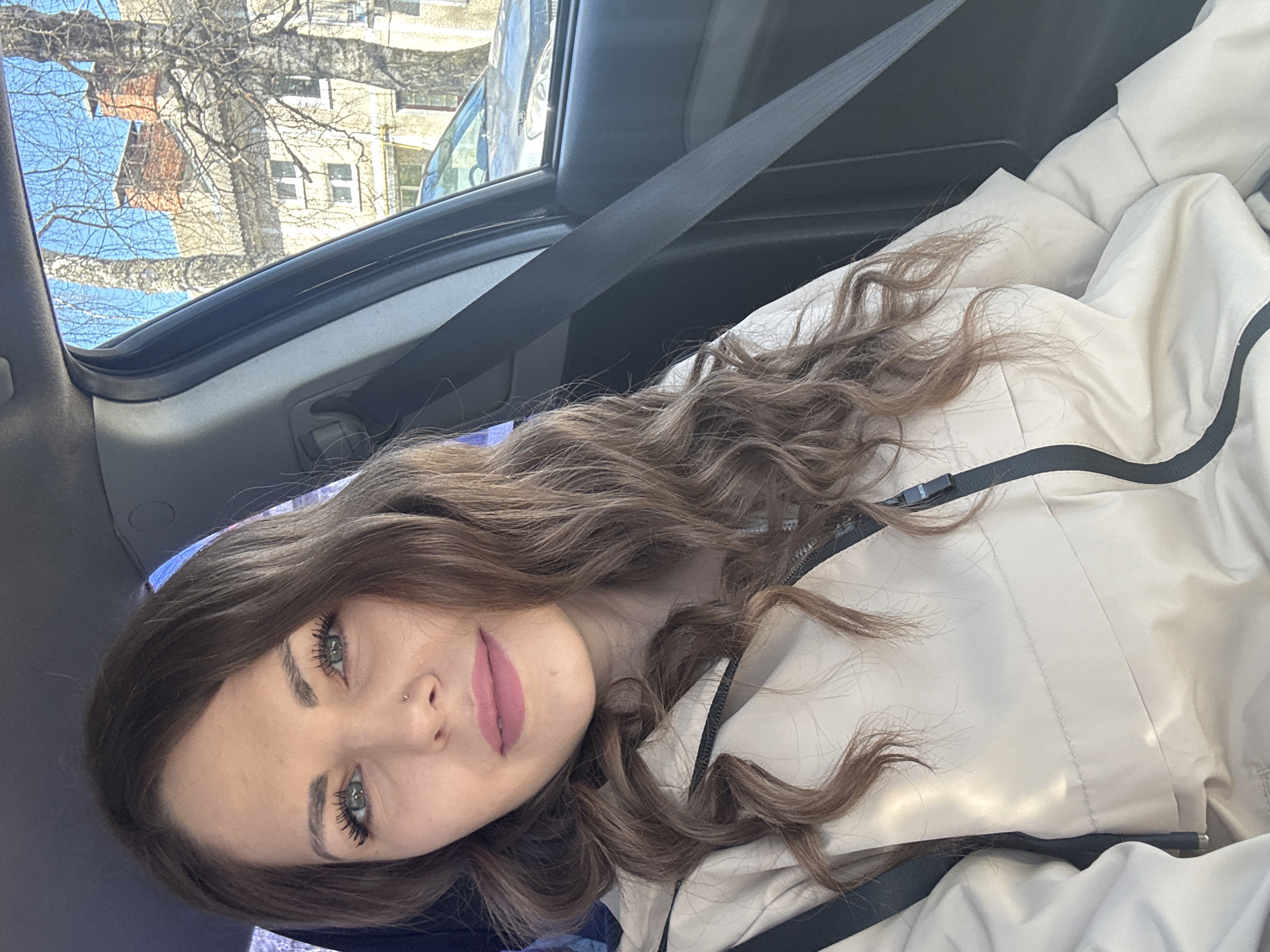 The photograph shows a woman sitting in the passenger seat of a car. She is wearing a seatbelt and a light-colored jacket. Her long, wavy brown hair is partially covering the jacket. The background visible through the car window includes a building and bare trees. The location is Раздолье, Russia, based on the buildings and vegetation.