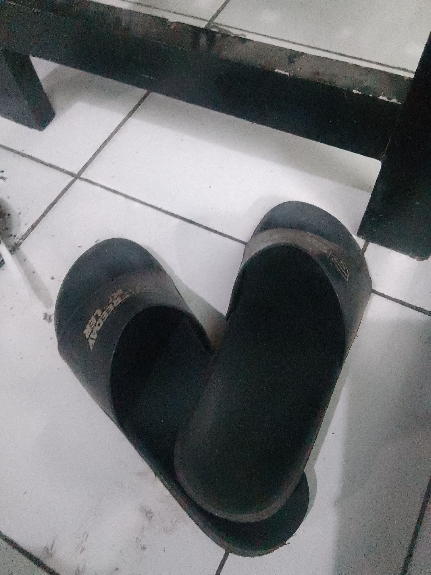 A pair of black rubber flip-flops are positioned on a white tiled floor. The flip-flops are placed side-by-side, with the left flip-flop oriented slightly behind and to the left of the right flip-flop, forming a heart-like shape. The left flip-flop has the word "FREEDAY" embossed on its strap in a white, stylized font. The right flip-flop appears to have a faint logo on its strap. A dark, wooden structure with a textured surface is visible in the upper portion of the image, casting a shadow on the floor. The environment appears to be indoors, likely a residential or common area, possibly in Trenggalek, Indonesia, given the context provided.