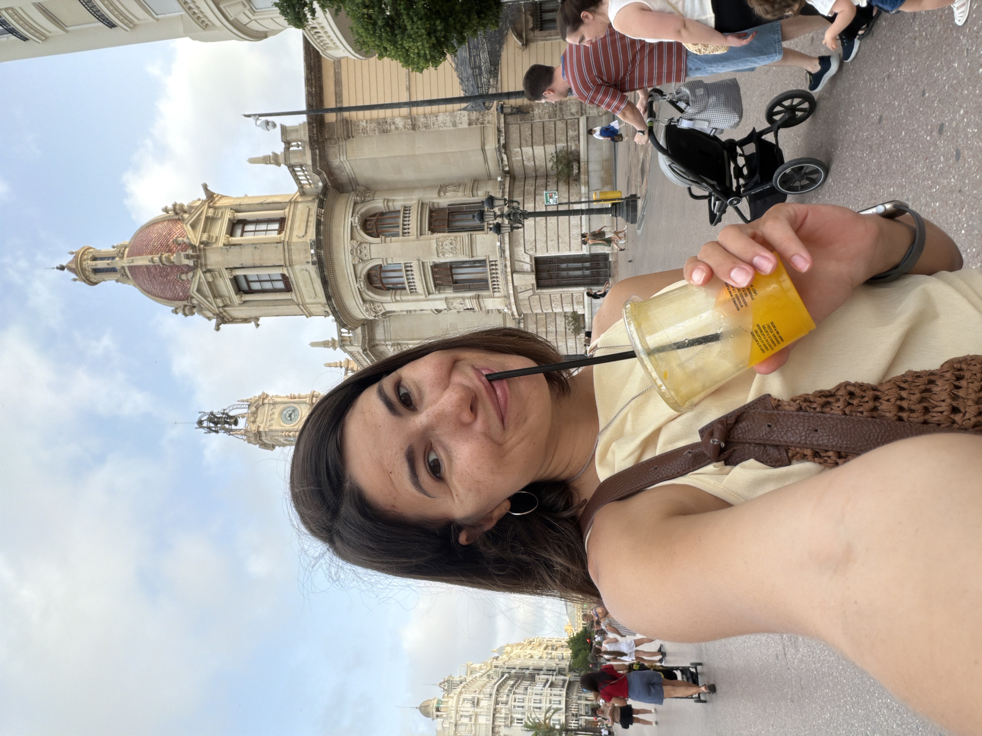 A medium shot captures a woman taking a selfie in an outdoor urban setting. She is positioned in the foreground, holding a clear plastic cup filled with an orange beverage and a black straw to her lips. Her gaze is directed at the camera, and she has a slight smile. The woman has long, dark brown hair and is wearing a light yellow top and a brown crocheted bag with a leather strap. Her left arm is visible, holding the drink.
In the background, a grand, ornate building with a dome and a clock tower stands prominently. The architecture suggests a European city. Several people are walking along a paved street in the middle ground. Some are casually dressed, and one person is pushing a stroller. The environment appears to be a public square or street, with the sky visible above, featuring scattered clouds. The lighting suggests it is daytime.