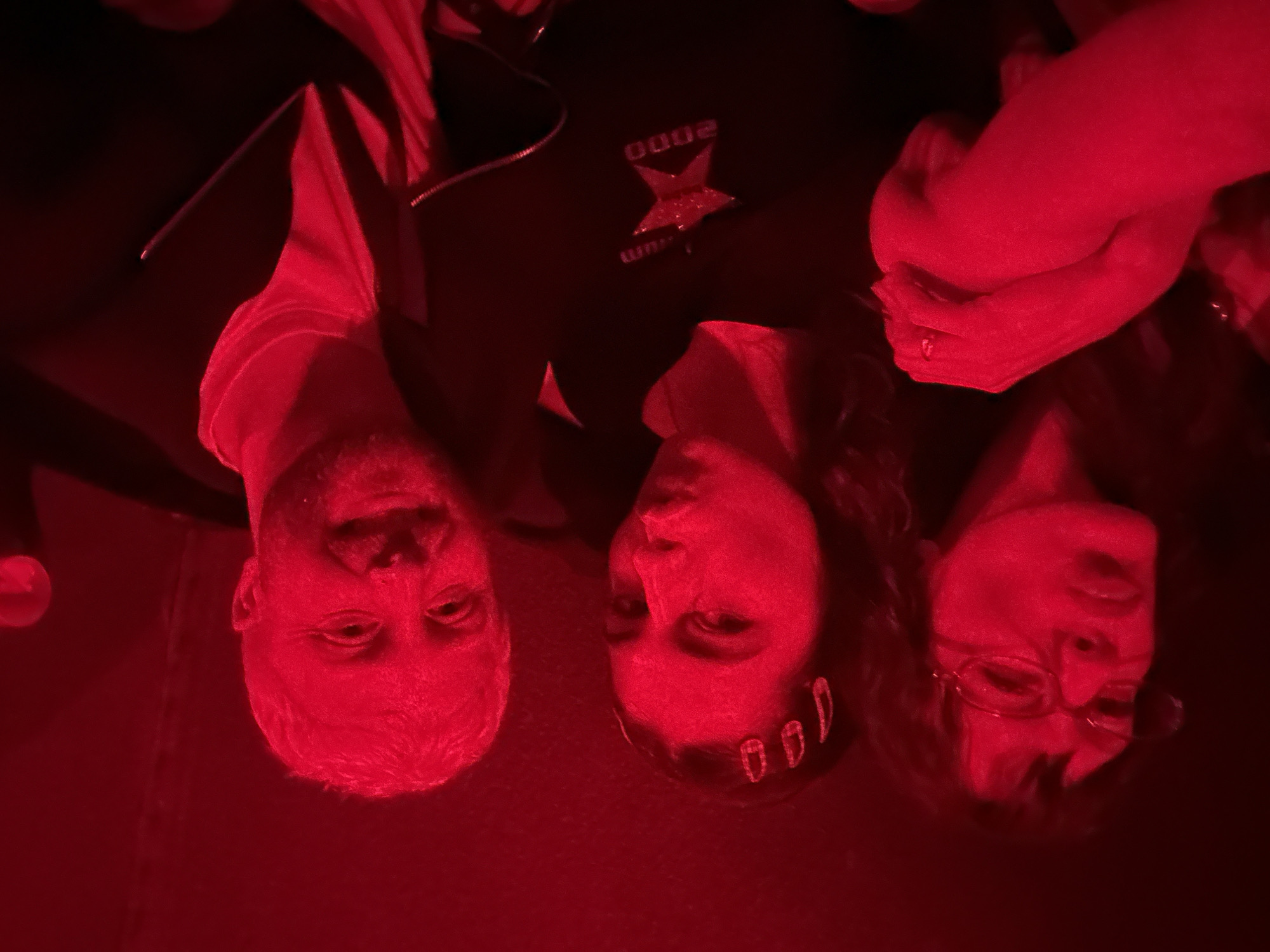 A close-up, rotated, harshly lit, red-toned image shows three people shoulder-to-shoulder. From left to right, a man with short, light hair and a short beard. The person in the middle is a woman with dark hair, wearing a hair accessory. The person on the right is a woman with dark hair, wearing glasses. The middle figure is wearing a black jacket with a graphic design that includes the number "2000." Some clasped hands are visible on the right. No location can be determined based on the visual information provided.