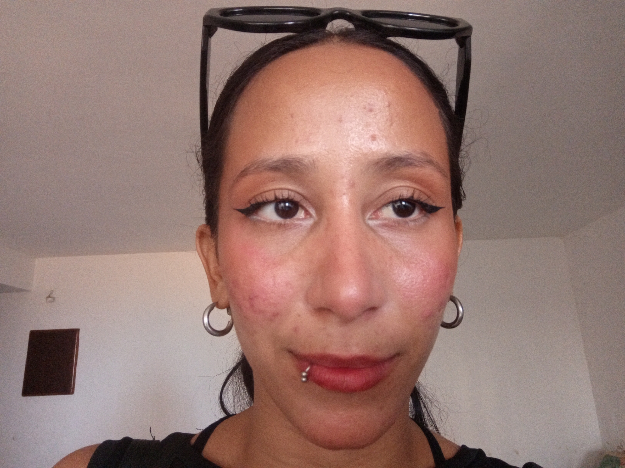 A close-up, frontal portrait of a person with tan skin. The person has black eyeliner on their upper eyelids and red lipstick. They have a lip piercing on the lower lip and silver hoop earrings. Black-framed sunglasses are perched on their head. The background is a plain, light-colored wall. A small, dark, square object is visible on the wall behind the person's left shoulder. The location, Porlamar, Venezuela, cannot be determined from the image alone.