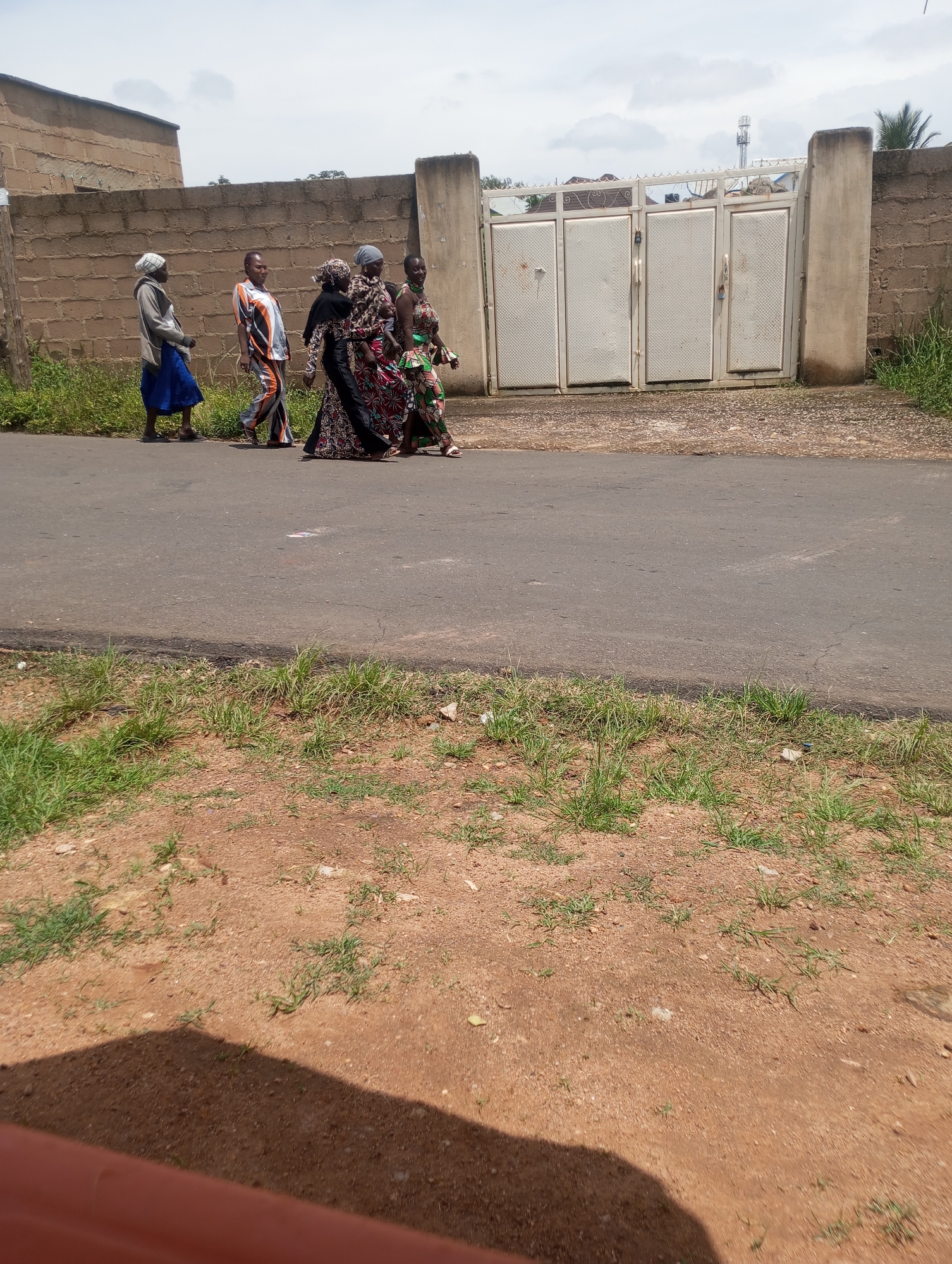 A group of five women are walking on a paved road, on the left side of the image. The woman in the front is wearing a light blue skirt and a grey hooded jacket. Next to her is a woman in a white shirt with orange and black stripes, and orange pants. Behind them, three women are dressed in brightly colored, patterned dresses and headscarves. All women appear to be walking in the same direction, away from the viewer.
In the background, a beige brick wall runs horizontally across the image, with a white metal gate in the center. The gate has four panels and appears to be closed. A cell phone tower is visible in the distance behind the gate. The road is paved and appears to be in good condition. The surrounding area is dry and dusty with patches of green grass growing along the side of the road. The sky is overcast with some clouds visible. The overall scene suggests a typical street in Jos, Nigeria, with people going about their daily activities.