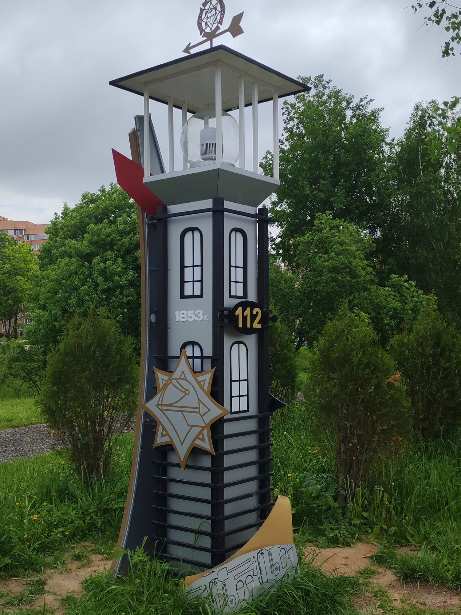 The image captures a stylized representation of a lighthouse located in an urban green space in the city of Unknown, Unknown. The structure is painted in grayscale, with black accents outlining window frames, horizontal bands, and support posts. It features white "windows" on each story, a gold star-shaped emblem, and the number "112" alongside the year "1853" in a gold font. Above, a small tower with a weather vane is affixed to the top. The surrounding environment includes verdant bushes and tall grasses, with trees and a building visible in the background under a cloudy sky.