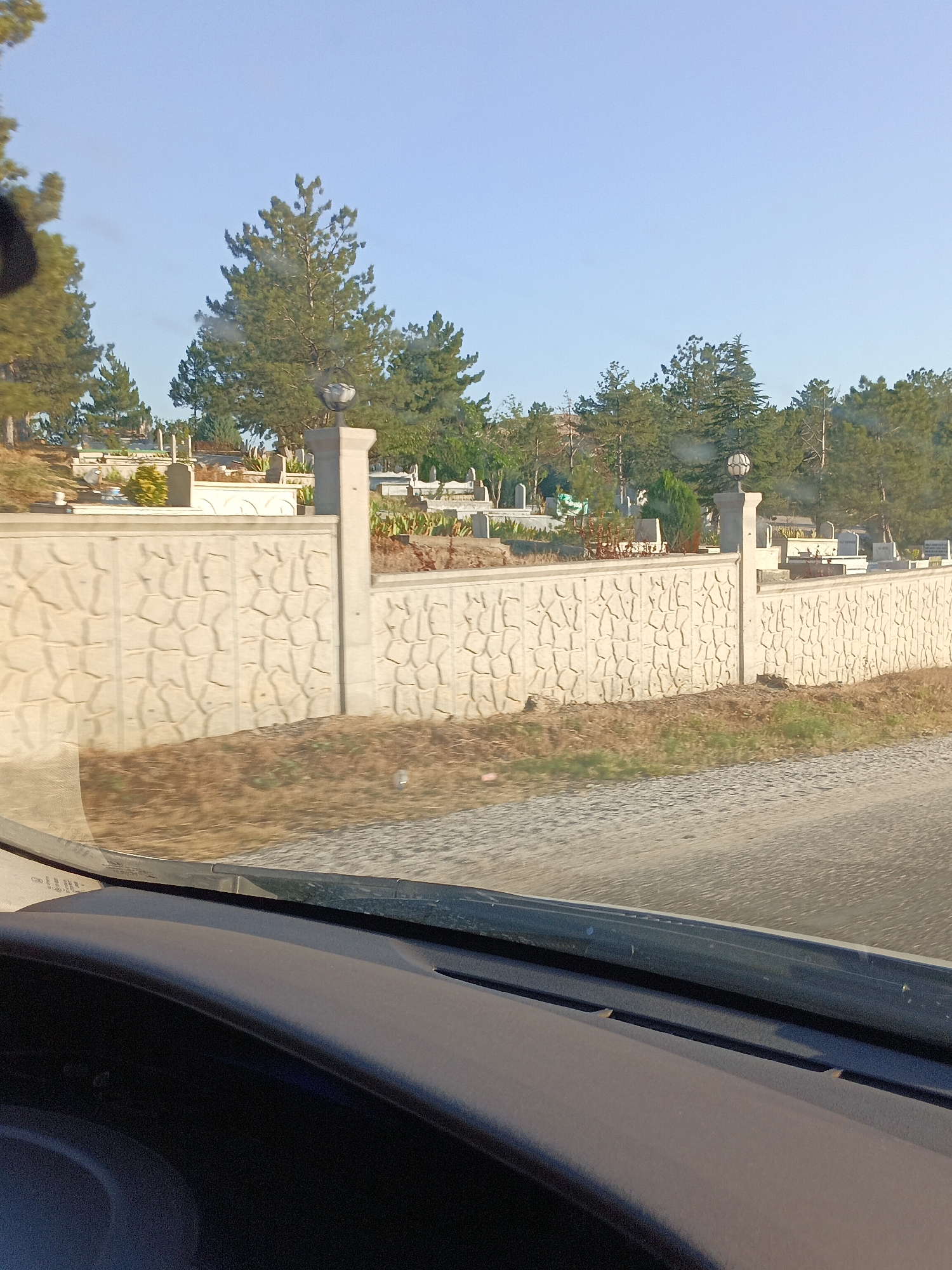 The image shows a view from inside a vehicle looking out at a cemetery. A textured concrete wall with decorative pillars and lamps separates the road from the cemetery grounds. The cemetery contains numerous gravestones and trees, with a clear blue sky overhead. The scene is likely in Bahşılı Belediyesi, Turkey, given the context of the prompt.