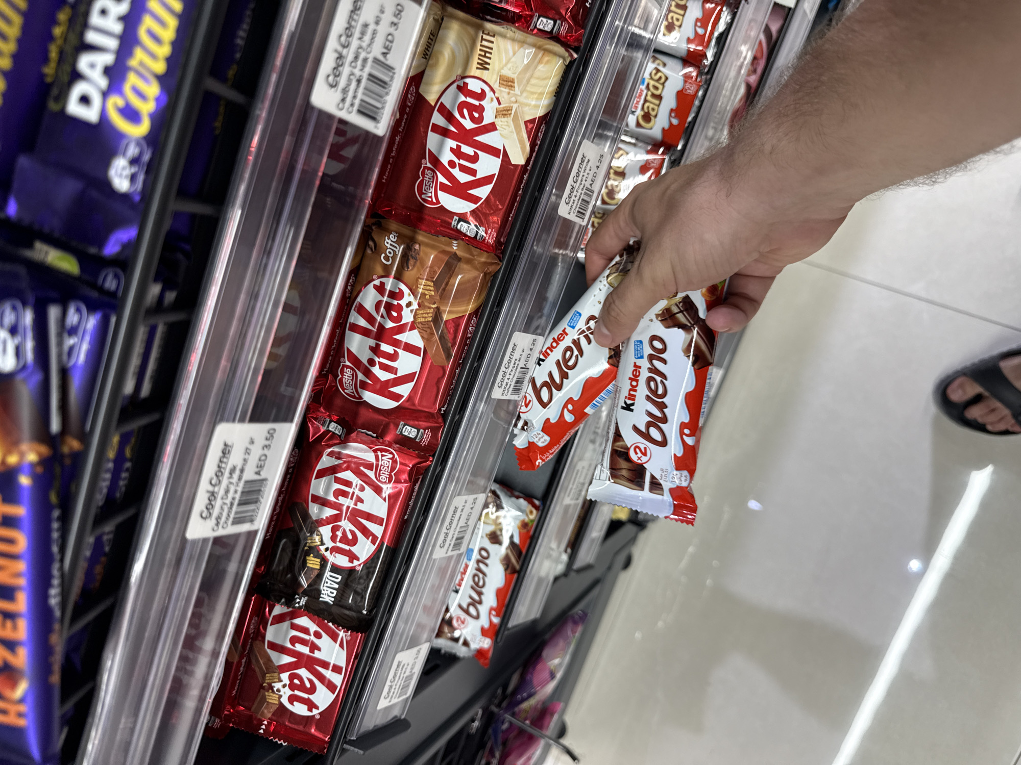 A person's hand is reaching for a candy bar on a store shelf in Dubai, United Arab Emirates. The shelf is stocked with various chocolate bars, including KitKat, Cadbury, and Kinder Bueno. The lighting is bright and the store appears to be a convenience store or supermarket. The person is wearing black sandals, and their foot is visible at the bottom of the frame. The shelves are made of metal and are organized with price tags. The candy bars are arranged neatly on the shelves.