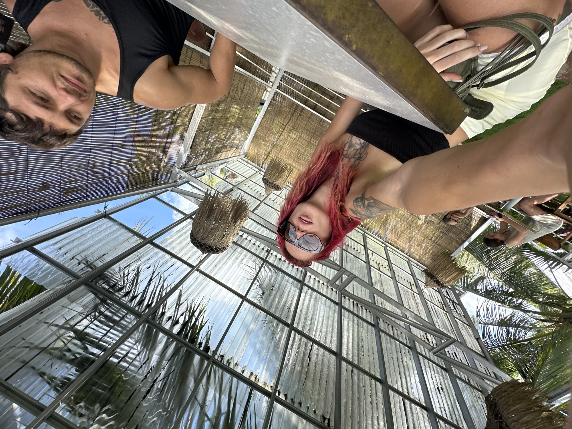 The photograph, taken in what appears to be a greenhouse structure on Nusa Penida, Indonesia, features a group of people around a metal-topped table.
One individual, a man, wears a black tank top and is positioned on the left. A woman with red hair, tattoos, and sunglasses is visible across the table. The structure's roof is made of clear or translucent panels with a metal frame. Exterior vegetation is visible through the roof and walls.
Other people can be seen in the background, further away from the camera's perspective.