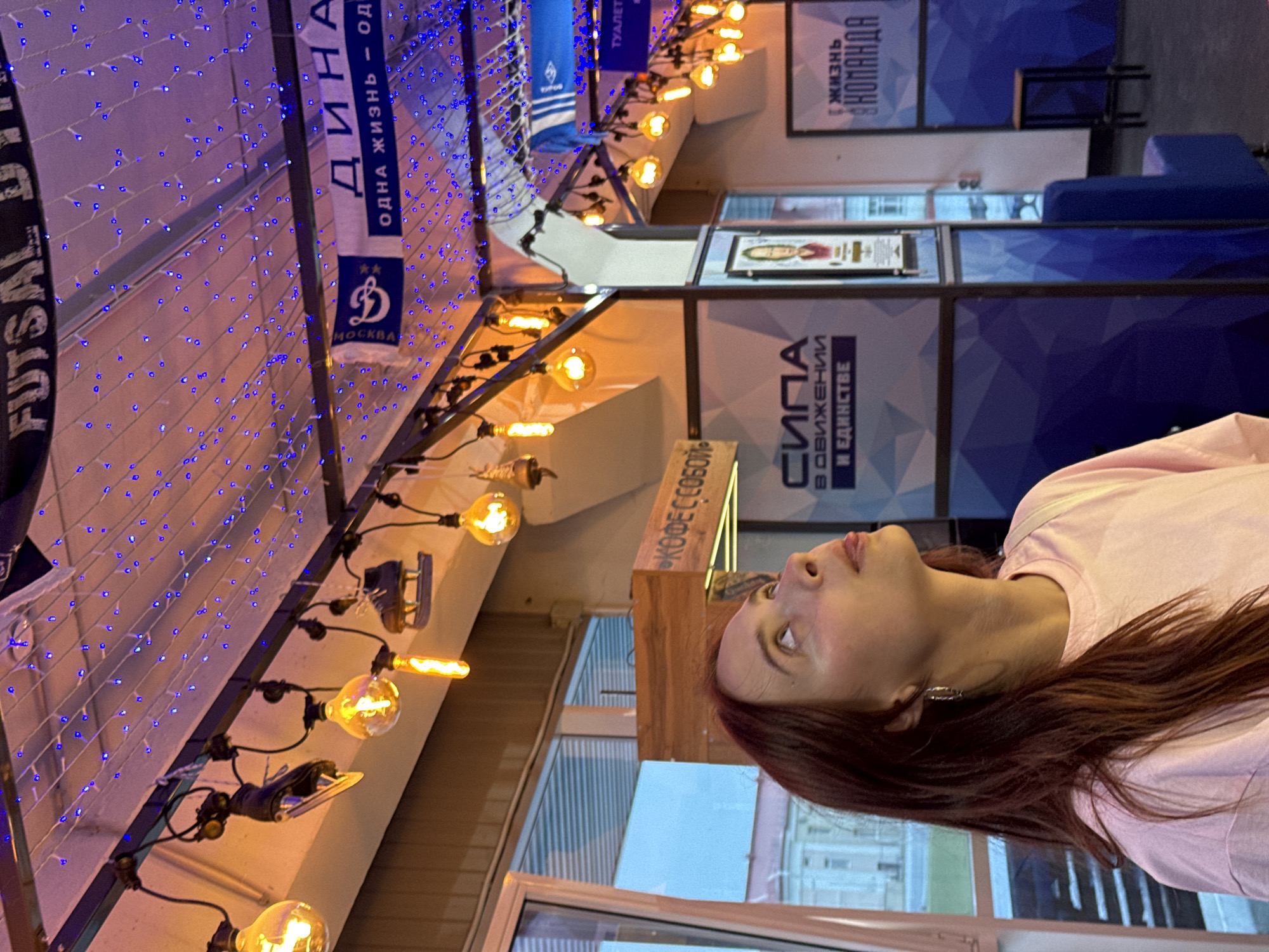 A woman with long, dark red hair and a light pink t-shirt looks up and to her right. She is positioned on the left side of the frame, with her head and shoulders visible. Her tongue is slightly sticking out. Behind her, an indoor space is decorated with strings of small blue lights and vintage-style Edison bulbs hanging from a metal frame. Several pairs of ice skates are also suspended from the frame. In the background, there are signs with Cyrillic text. One sign reads "ДИНАМО" and another appears to say "КОФЕСОБОЙ". The overall environment suggests a recreational facility or an event space, possibly a cafe or sports-related venue in Izhevsk, Russia.