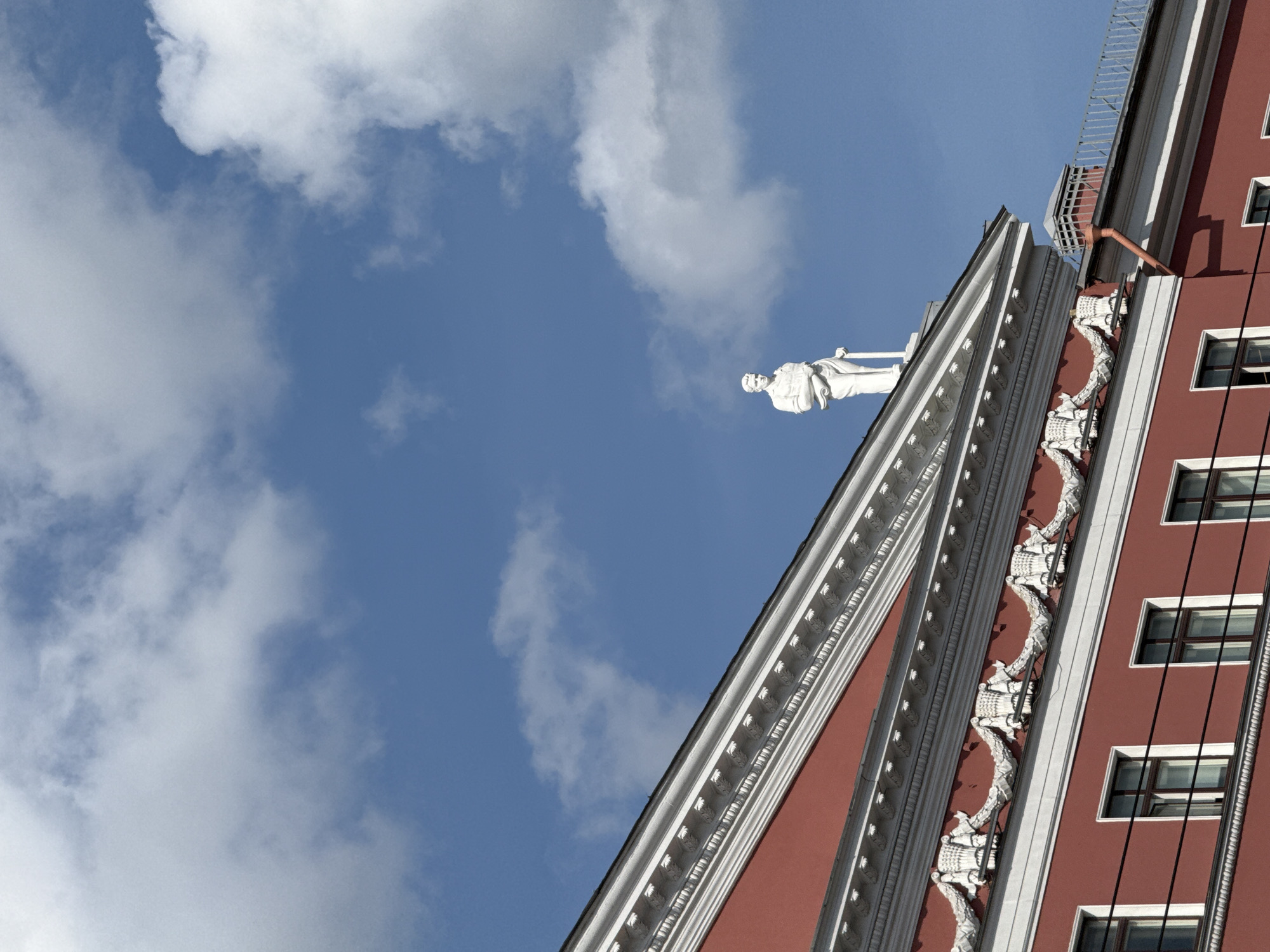 The photograph depicts a fragment of a building facade against a backdrop of a blue sky with scattered white clouds. The building is red with white architectural details, including decorative moldings and window frames. A white statue of a figure is positioned on the roofline. The building is located in Saint Petersburg, Russia, and is identified as the apartment building of the insurance company "Russia." The view is angled upwards, emphasizing the building's height and the architectural details.