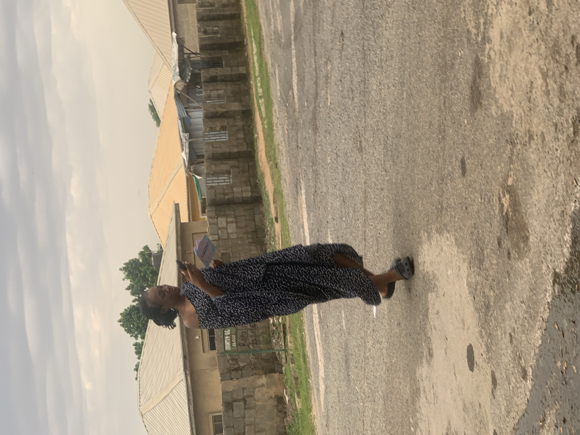 In Abuja, Nigeria, a young woman in a dark, off-the-shoulder dress with a white speckled pattern is walking on a paved road. She is holding a mobile phone in her right hand and a small, light-colored object, possibly a card or a pamphlet, in her left hand. She appears to be looking down at these items. Behind her, there is a low, grey stone wall and beyond that, a series of houses with corrugated metal roofs in varying shades of yellow and grey. A green signpost is partially visible near the wall. The sky is overcast with grey clouds. The road surface is asphalt with some patches of lighter colored concrete and evidence of puddles.