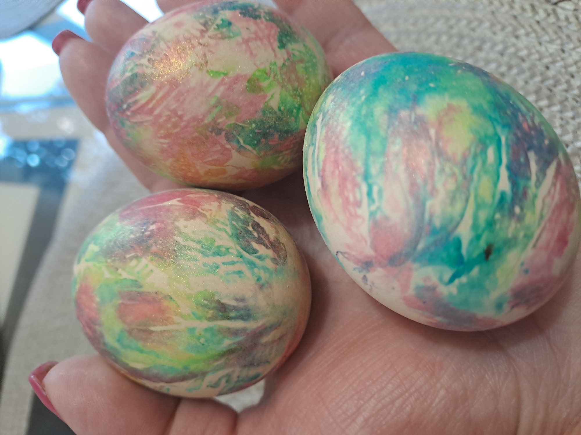 A close-up shows a person's hand holding three decorated Easter eggs. The eggs have a marbled effect with colors including pink, green, blue, and a base color of cream. The person's fingernails are painted a matching shade of pink. The background is blurry, but appears to be a neutral-colored surface. The location, Elektrostal, Russia, cannot be determined from the visual content of the image.