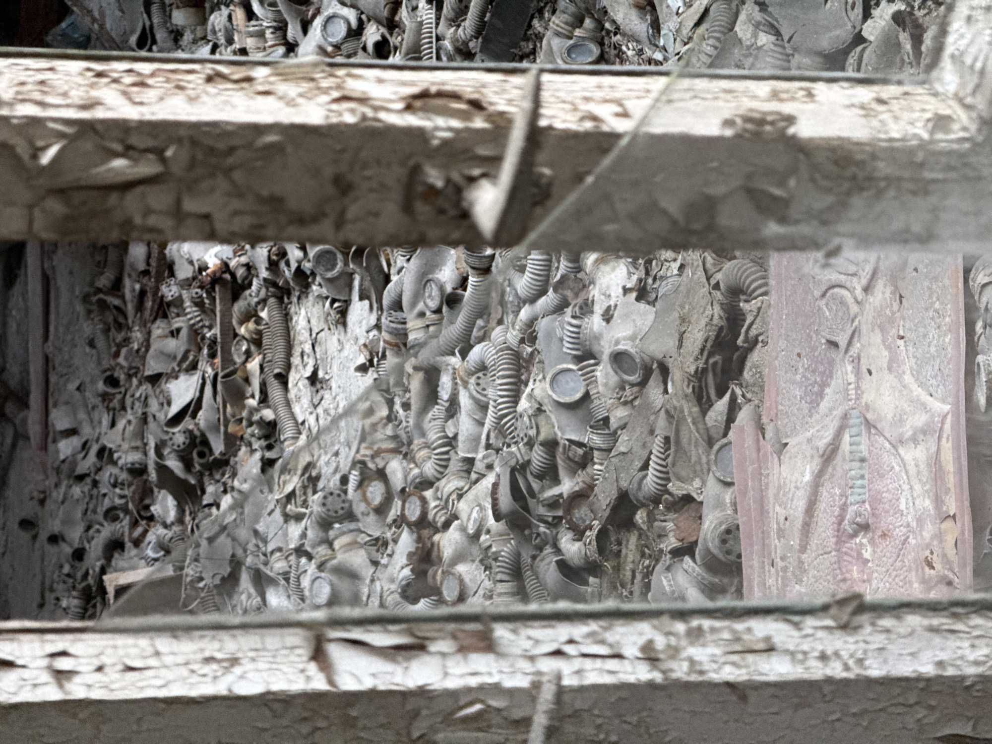 The image shows a view through a broken, decayed window frame filled with peeling paint. The foreground consists of the window structure itself, with a shattered pane still partially in place. The scene beyond is dominated by a chaotic pile of discarded gas masks and their associated corrugated hoses. The masks appear to be uniformly grey and dust-covered, adding to the impression of abandonment and decay. The setting suggests the location is within the abandoned city of Pripyat, Ukraine, near the Chernobyl Exclusion Zone.