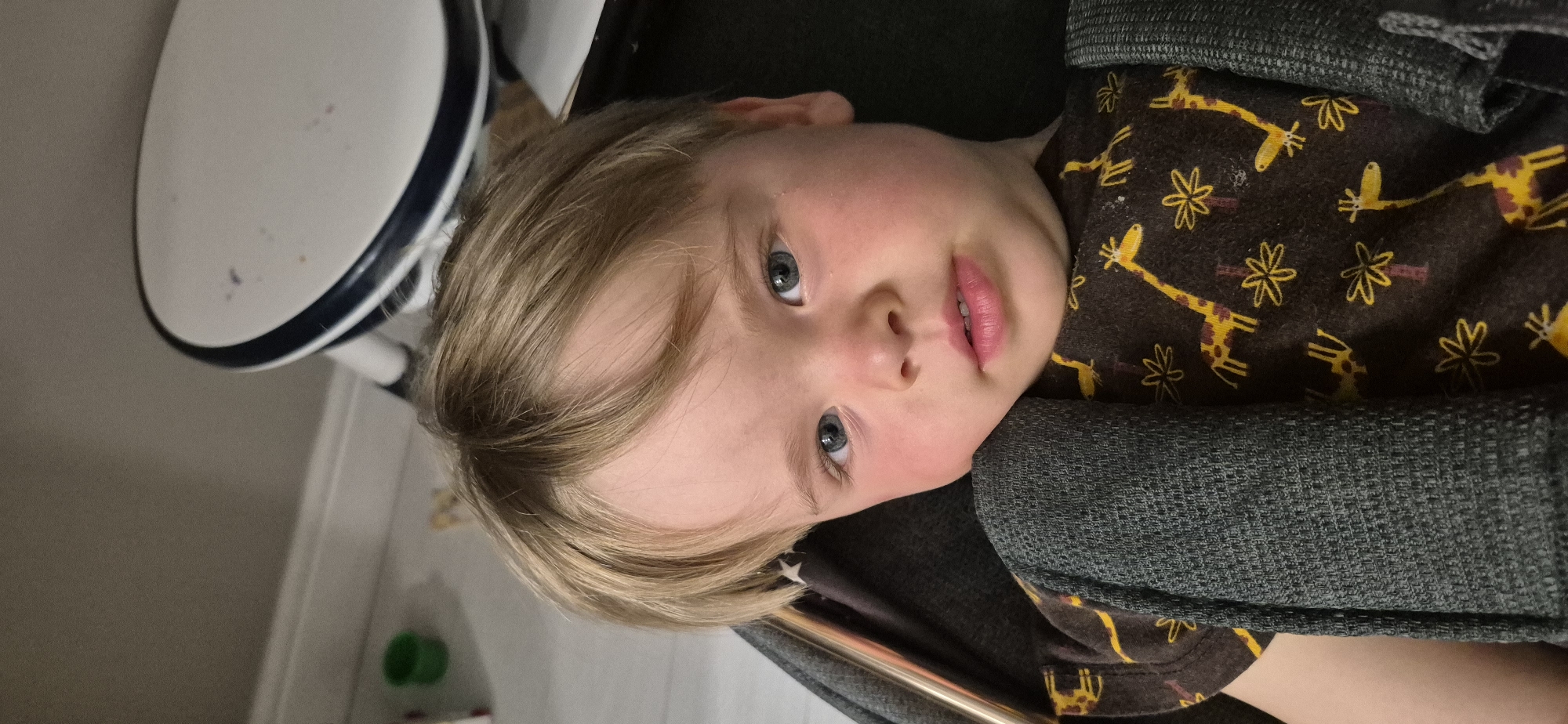 A close-up, eye-level, vertical medium shot shows a young boy with blonde hair and blue eyes. He is wearing a dark brown shirt with a pattern of yellow giraffes and flowers. A dark gray textured blanket is draped over his shoulder and chest. His face is turned slightly towards the camera, and his cheeks are flushed pink. His lips are slightly parted. In the blurred background, a portion of a white and blue object, possibly a chair or table, is visible. To the left of the boy's head, in the upper left corner of the image, a small green plastic cup is visible on a light-colored floor. The overall impression is that of a child resting or being held.