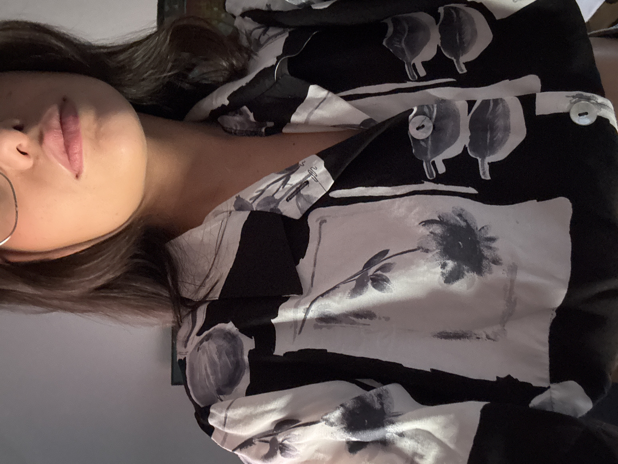 A close-up, head-and-shoulders shot of a young woman wearing a black and white floral print button-down shirt is depicted. She is wearing thin, wire-rimmed glasses and has dark brown hair. The background is a neutral gray wall. The image is taken from a medium-close perspective, focusing on her upper body and face. The lighting appears to be artificial, creating soft shadows. The scene does not provide any information that could be used to determine the location in Las Rozas de Madrid, Spain.