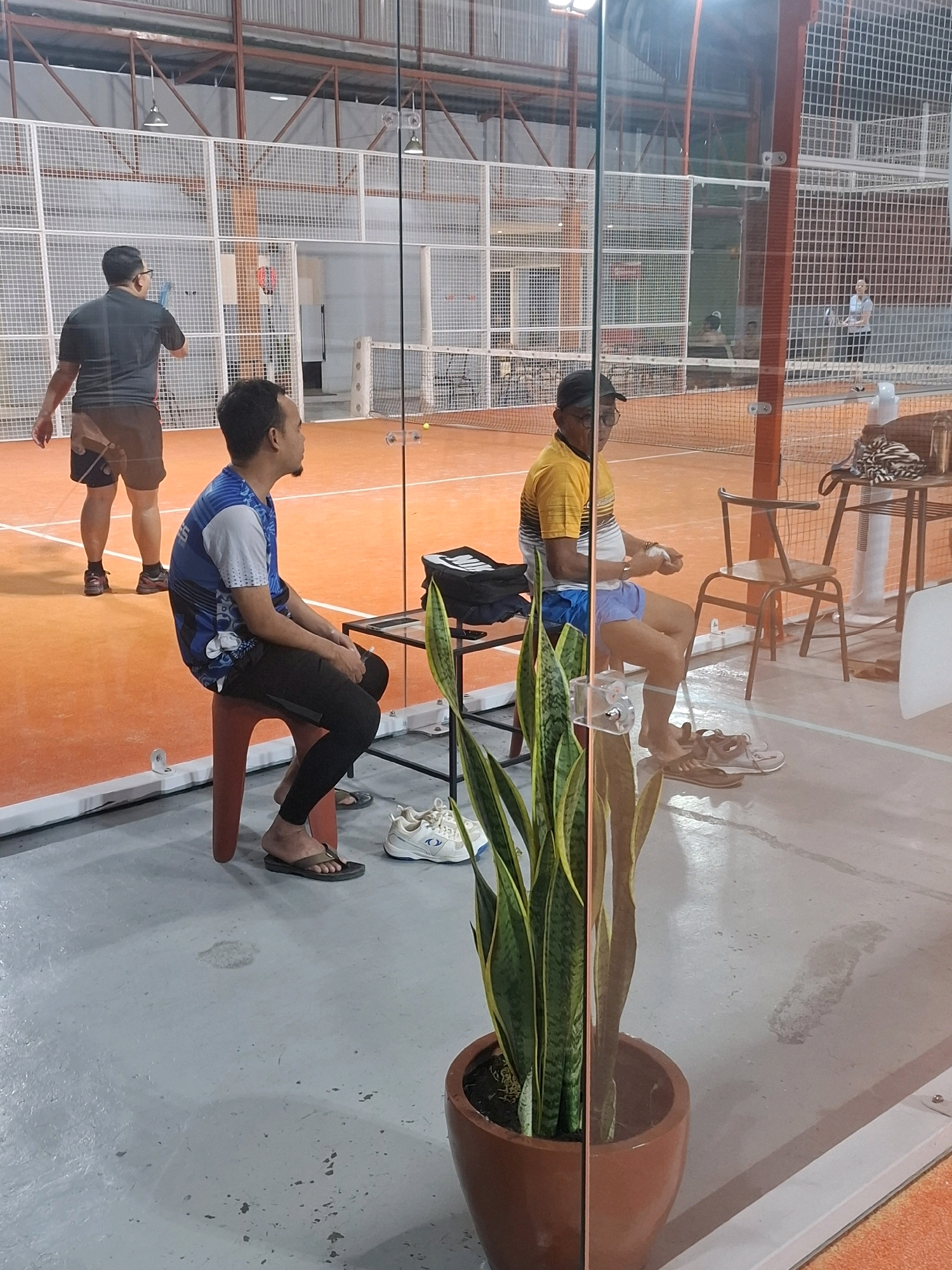 In the city of Cisaat, Indonesia, an indoor sports facility, likely a padel court, is depicted. The court features an orange playing surface and is enclosed by glass walls and white netting. Two men are prominently visible. One man, wearing a dark t-shirt and shorts, stands on the court with a racket in his hand, facing away from the camera. Another man, wearing a blue and white patterned t-shirt and black leggings, sits on a red stool on the sideline. He is looking to his right and appears to be resting. A third man, wearing a yellow and white striped t-shirt and blue shorts, is seated on a chair in the background, holding a white object, possibly a ball, and looking forward. Several chairs and tables are also present around the court. A potted snake plant is in the foreground, partially obscuring the view of the scene. The overall lighting suggests an indoor setting, possibly illuminated by artificial lights.