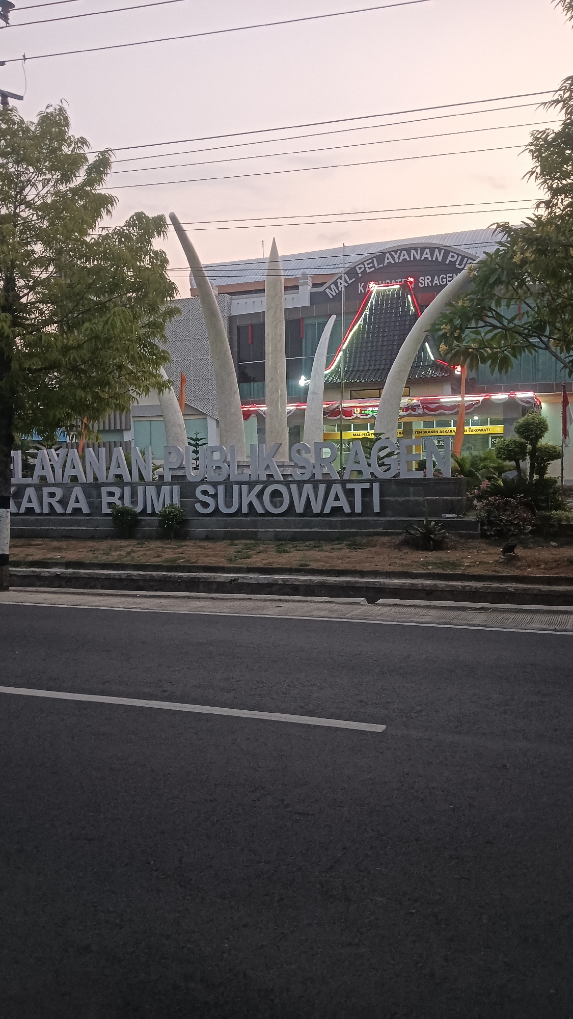 This is a vertical, eye-level shot of the front of the Public Service Mall of Sragen in Sragen Kulon, Indonesia, with a street in the foreground. The sky is a pale pink and orange, suggesting dusk or dawn. In the foreground is a dark asphalt road with a single white line marking the lane. Behind the road is a raised area with a concrete border, then a well-maintained grassy area with a few small bushes. Behind the grass is a large, raised, three-dimensional sign with white block letters that spell out "LAYANAN PUBLIK SRAGEN KABUPATEN SUKOWATI." The letters are illuminated by the ambient light and appear to be made of stone or concrete. Behind the sign are four tall, white, abstract sculptures resembling elongated leaves or flames.
The building behind the sculptures is the Public Service Mall of Sragen. It is a modern building with a gray, patterned facade and a dark roofline. Some of the windows are visible, and there are red and white banners hanging across the front of the building. Above the entrance, a sign reads "MAL PELAYANAN PUBLIK KABUPATEN SRAGEN" in large letters. There are also smaller signs and what appear to be flags visible. To the right of the building, there are some trees and manicured bushes. The overall scene conveys a sense of civic pride and the provision of public services.