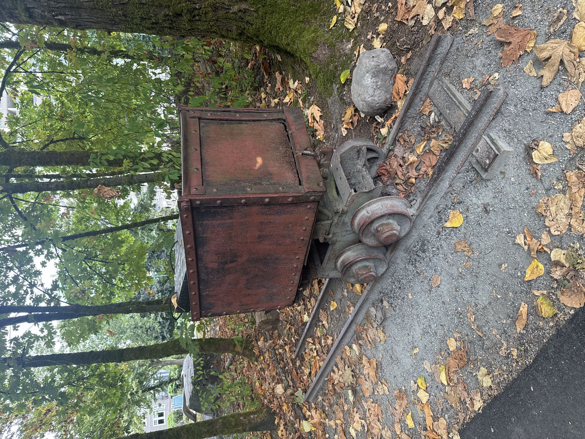 An abandoned, rust-colored mine cart sits on railway tracks in a wooded area. The cart is made of metal and appears to be old and weathered. The tracks are made of metal and are partially covered in fallen leaves. The area is surrounded by trees and foliage, and there is a large rock to the right of the cart. In the background, buildings can be seen, suggesting an urban or suburban setting. The image was taken outdoors, likely during the autumn, given the presence of fallen leaves. The scene is static and depicts a historical artifact in a natural setting.
This scene is located at the Fraser River Discovery Centre in Coquitlam, Canada. The mine cart is a relic from the local mining history, specifically from the historical coal mining operations that were once prevalent in the region. The Fraser River Discovery Centre is a park that preserves and showcases various historical artifacts related to the area's industrial past.
