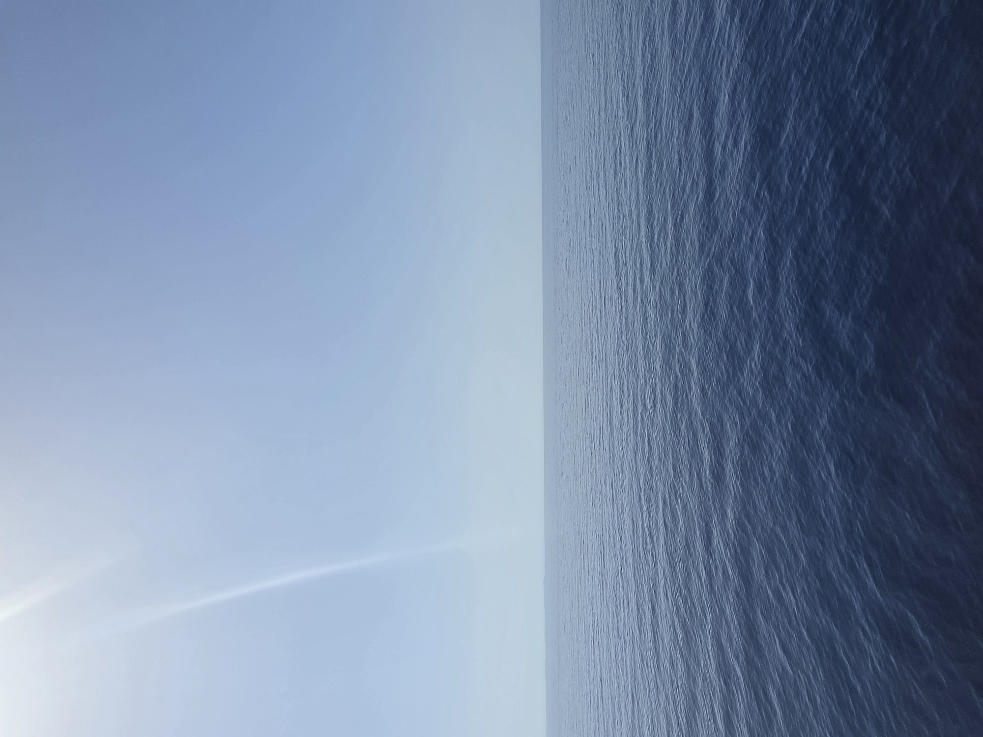 The image shows a wide expanse of calm, dark blue ocean under a clear, pale blue sky. The horizon line is sharp, dividing the water from the sky. A faint, bright streak of light, possibly a sunbeam or lens flare, is visible in the upper left quadrant of the sky. The water's surface is gently rippled, indicating minimal wave activity. There are no people, boats, or landmasses visible in the frame, suggesting an open sea environment. The scene is devoid of any urban elements or specific landmarks that would place it within the city of Santa Ponça, Spain.