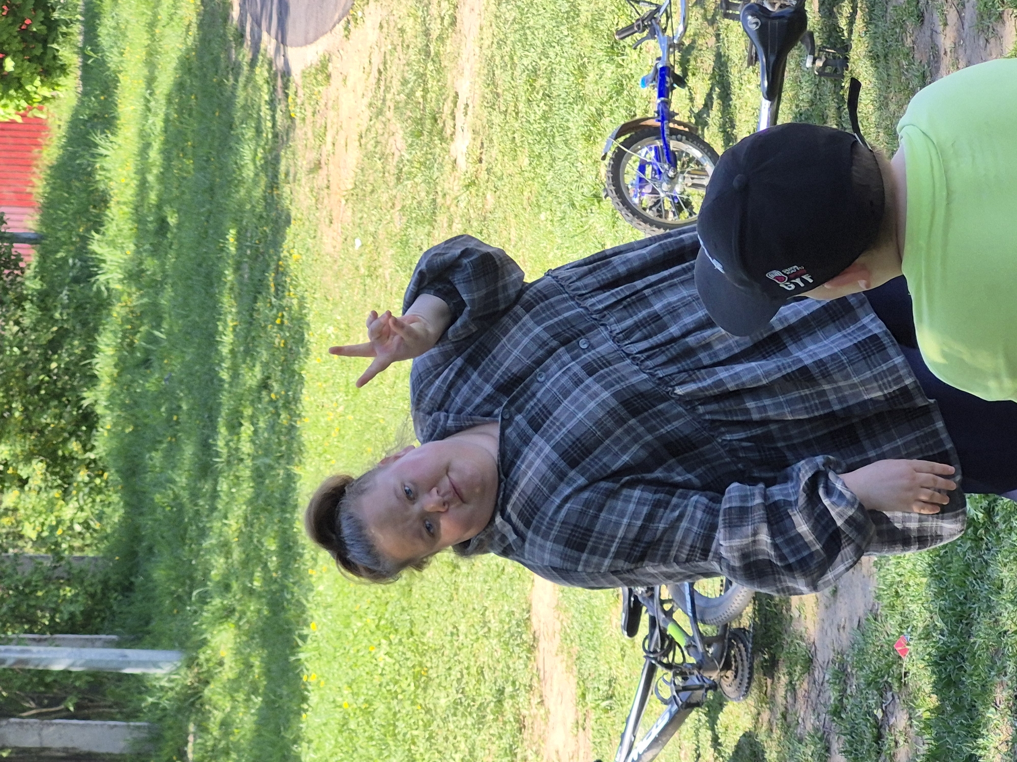 The image depicts a woman in a plaid shirt standing on grass, giving a peace sign with her left hand. To her right is a child wearing a green shirt and a black baseball cap. Two bicycles are nearby, one blue and one gray. The background features a grassy area with shadows from trees, and a fence is visible in the distance. The location appears to be an outdoor area with grass and trees. The city of Unknown, Unknown cannot be determined from the image.