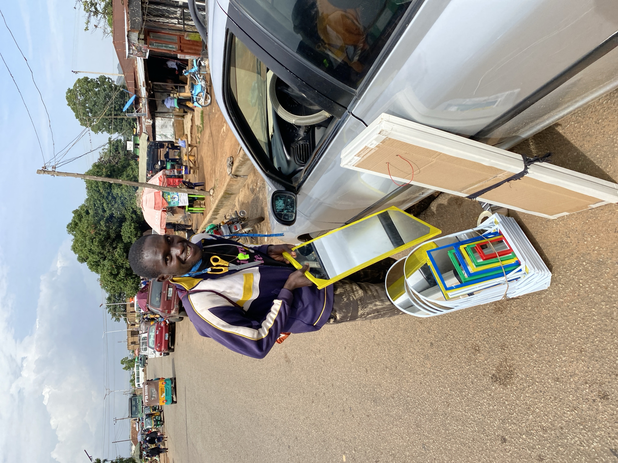 A man stands next to a silver car on a paved road, holding a rectangular mirror with a yellow frame. He is wearing a purple and yellow jacket and appears to be smiling. To his right, stacked on the ground, are several other mirrors of various sizes, some with colored frames, as well as flat, rectangular objects wrapped in cardboard and tied with string.
In the background, there is a bustling street scene in Bukuru, Nigeria. A roadside market with stalls and a red umbrella is visible, along with several vehicles including cars, a blue motorcycle, and colorful auto-rickshaws, locally known as "Keke Napep". Trees line the street, and utility poles with wires are present. The sky is partly cloudy, suggesting it is daytime. The overall impression is of a vibrant, commercial environment.