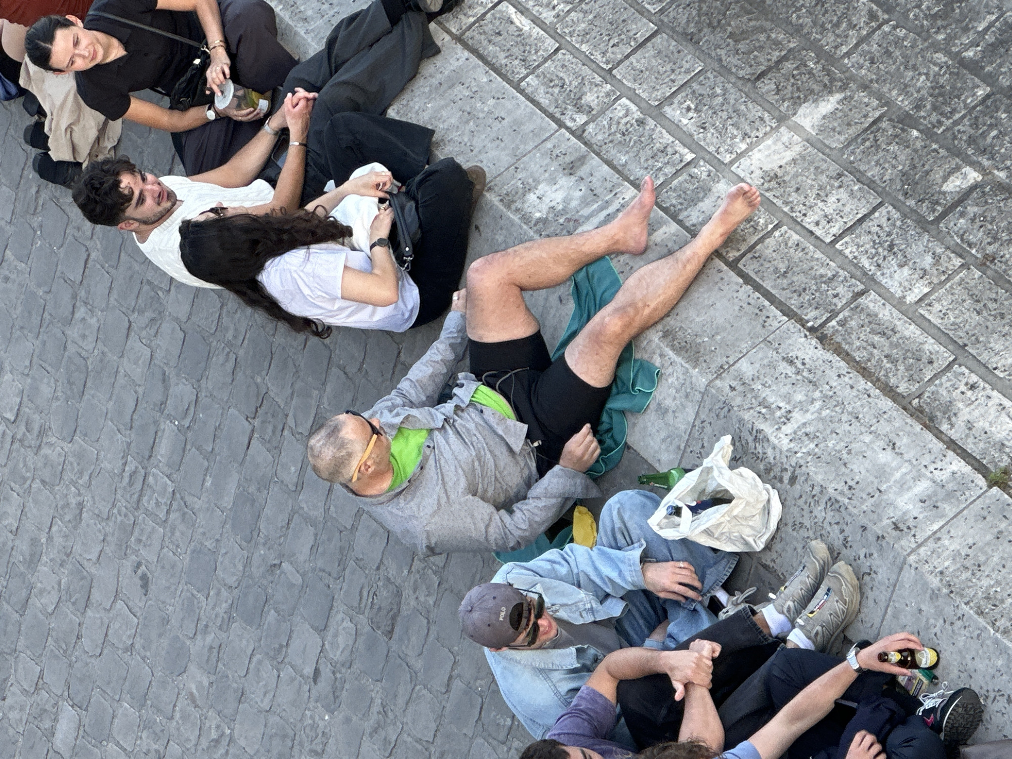 Several people are sitting on a brick surface, likely a public space in Paris, France. The image is taken from an overhead perspective. The group consists of what appears to be six individuals in casual wear, with some lying down while others are seated. Some are holding hands. There are several bottles and a white plastic bag present. The environment is an outdoor setting, with grey brick paving and a small stone wall.