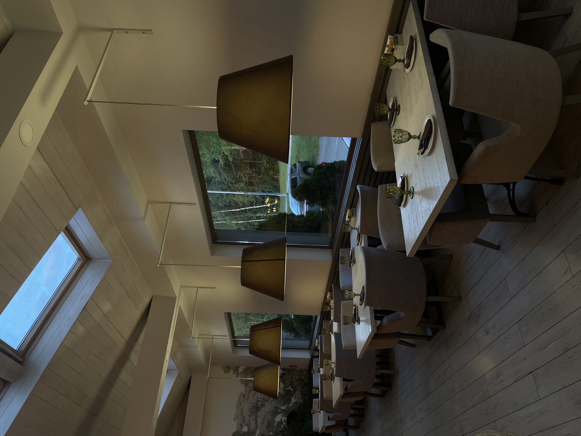 The image depicts the interior of a restaurant in Krasnogorsk, Russia, during twilight. Large windows offer a view of trees and a road with parked cars. The dining area is set up with multiple tables, each covered with light-colored tablecloths and adorned with green glassware and dark napkins. Upholstered chairs in neutral tones surround the tables. Overhead, large, dark lamps hang from the ceiling, providing ambient light. The ceiling features wooden beams and a skylight, through which a pale blue sky is visible. The floor is laid with light-colored wooden planks. A mural or wallpaper with a forest motif is visible on the left wall.
