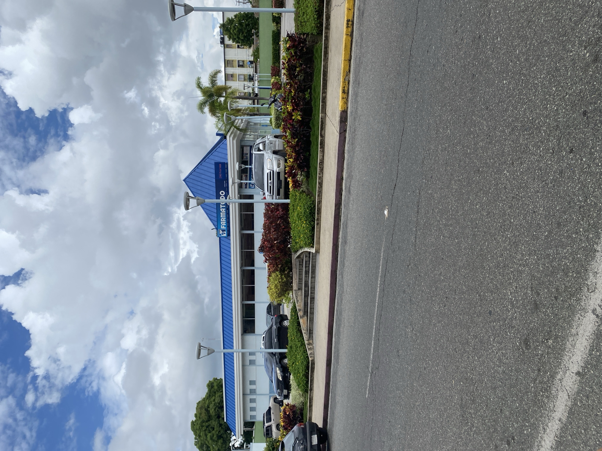 The image shows the Farmatodo store in Tinaquillo, Venezuela. The building is two stories with a blue roof and white walls. The Farmatodo logo is visible on the front of the building. Several cars are parked in front of the store. A street runs along the side of the building. The sky is cloudy. There are plants and streetlights along the sidewalk. A multi-story building is visible in the distance.