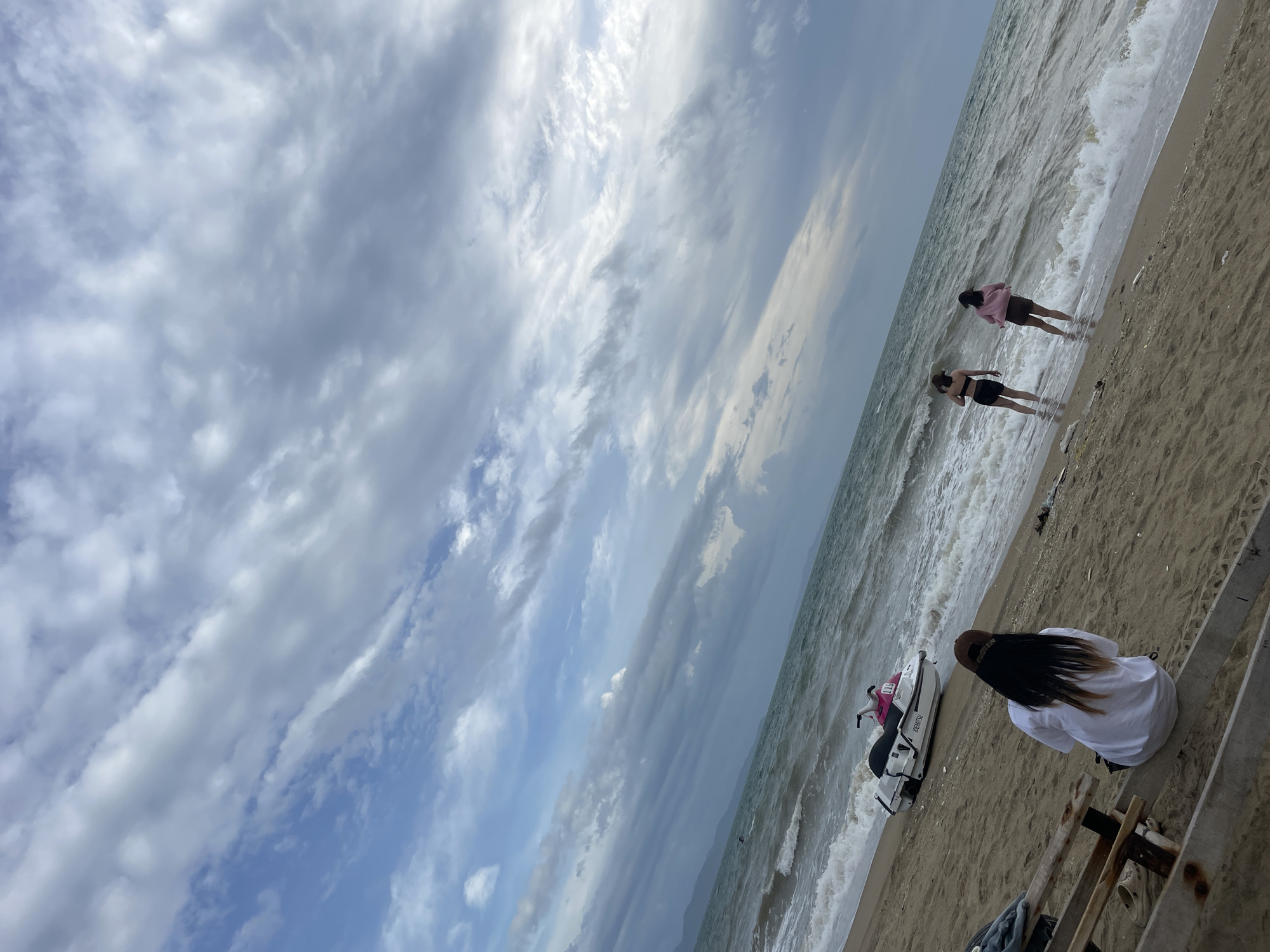 The image depicts a beach scene in Pattaya City, Thailand. In the foreground, a person with long dark hair and a white shirt is seated on a wooden structure, facing away from the camera and towards the ocean. Further down the beach, two individuals, appearing to be women, stand in the shallow water at the edge of the sea. One is wearing a black two-piece swimsuit, and the other is in a pink top and brown shorts. A white jet ski with pink accents is parked on the sand near the water's edge. The sea is choppy with visible waves crashing onto the shore. The sky is filled with dramatic, layered clouds, suggesting overcast or approaching weather. The background reveals a distant coastline with mountainous terrain. The overall environment is a sandy beach with the ocean and a cloudy sky.