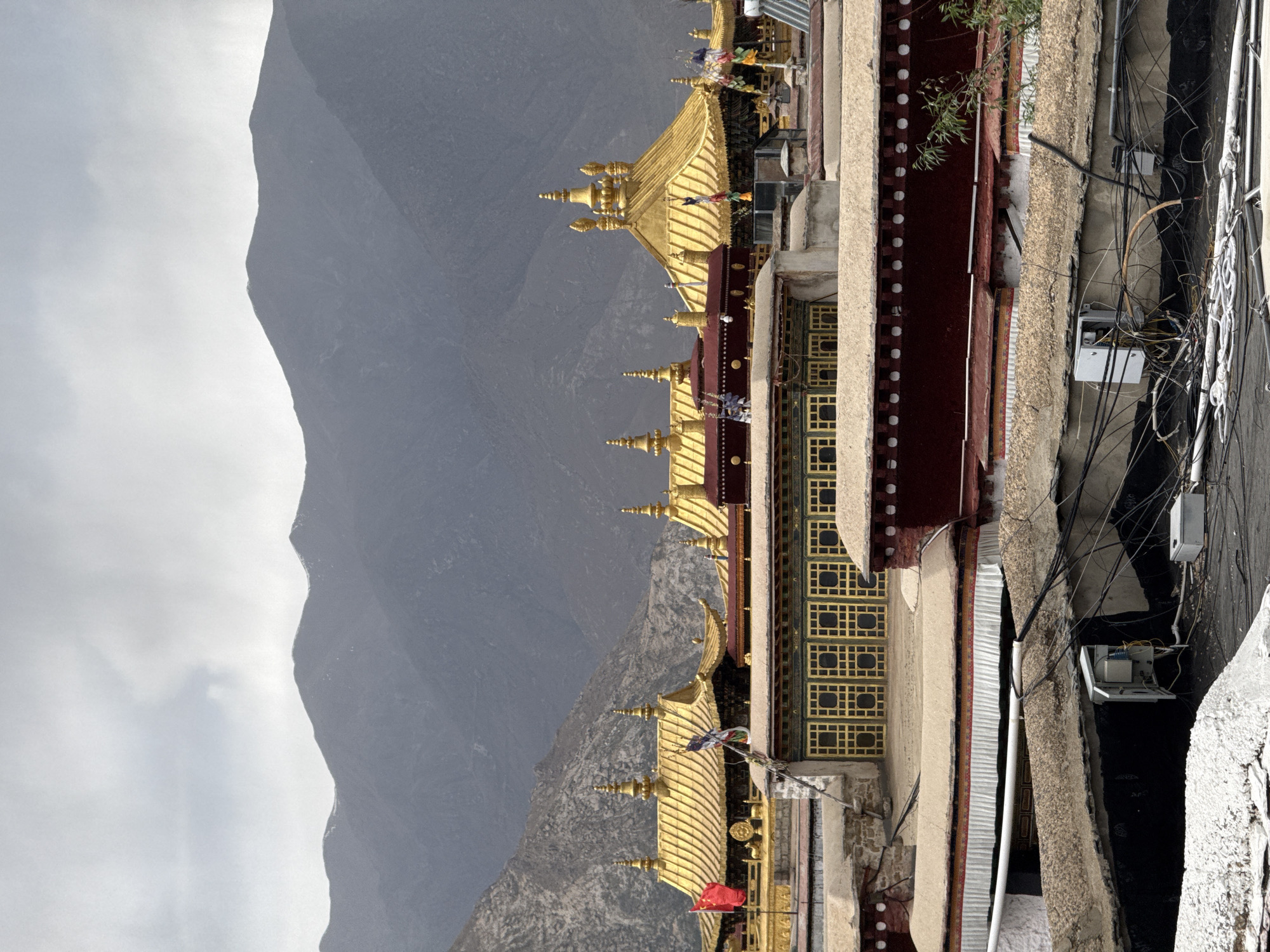 The image captures a building with a golden roof against a backdrop of a mountain range in Chengguan District, Lhasa, China. The building has red and beige walls, with a series of ornate golden roof structures. The mountain range appears grey and imposing. The foreground includes a street with electrical wires and utility boxes.