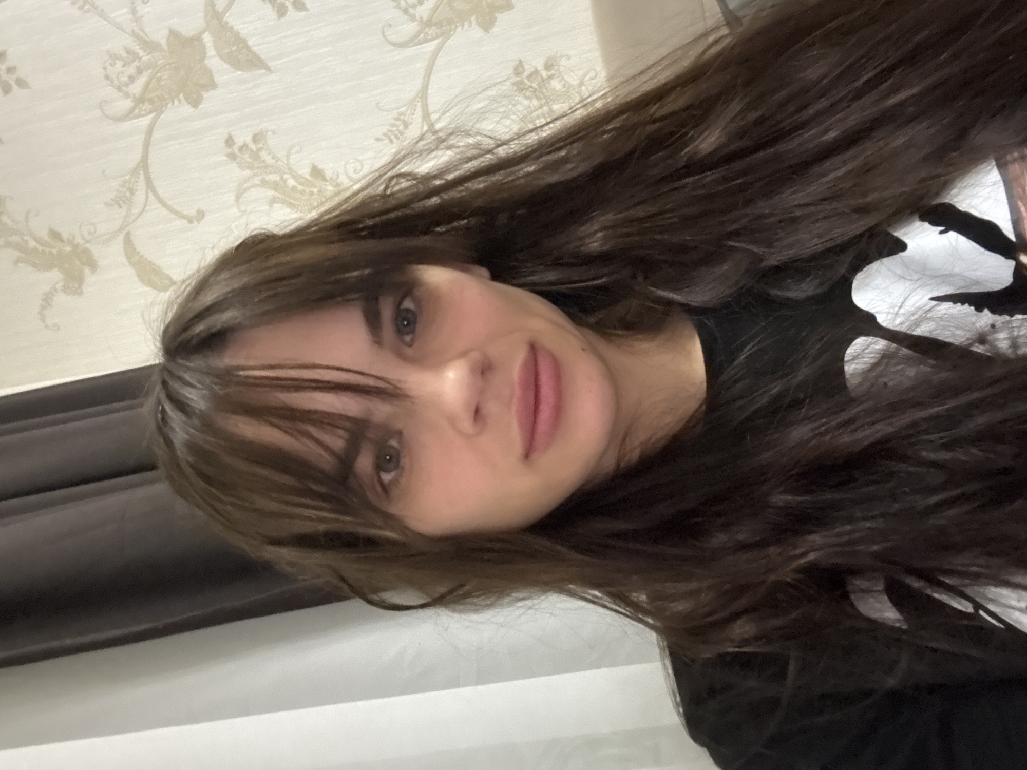 A young woman with long, dark brown hair and bangs is featured in a close-up indoor portrait, looking directly at the camera. Her head is tilted, revealing light-colored (likely green or hazel) eyes and a soft, subtle smile on her lips. She has fair skin and is wearing a dark, possibly black, top or t-shirt. The background consists of light-colored wallpaper decorated with a delicate, ornate floral or vine pattern in a slightly darker, muted tone. To the side, a dark horizontal element, possibly a piece of furniture like a headboard, is visible, alongside a lighter surface. The lighting is ambient, indicating an indoor setting, likely in Rostov-on-Don, Russia. No visible text is present.