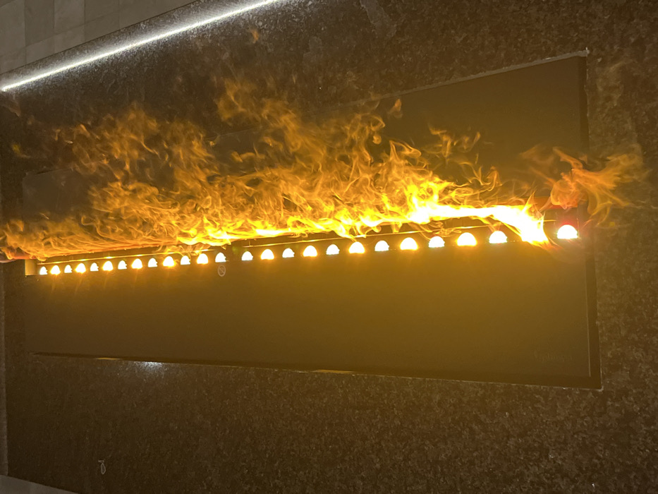 The image depicts a contemporary linear architectural fire and light feature, situated within the Paradise Center in Sofia, Bulgaria. The feature is set into a dark, speckled floor, likely granite. It comprises a long, narrow, recessed trough. Along the left side of this trough, a precise row of evenly spaced, half-dome-shaped LED lights emits a consistent, warm golden-yellow illumination. Adjacent to these lights, on the right side of the trough, vibrant orange and yellow flames actively burn and undulate, rising from an unseen source to form a dynamic "river of fire." The surrounding floor extends outward, presenting a dark, granular texture. On the far right, a linear strip of white ambient lighting is visible, embedded into an adjacent floor section or wall base. No individuals, overt actions, or interactions are visible within the frame.
