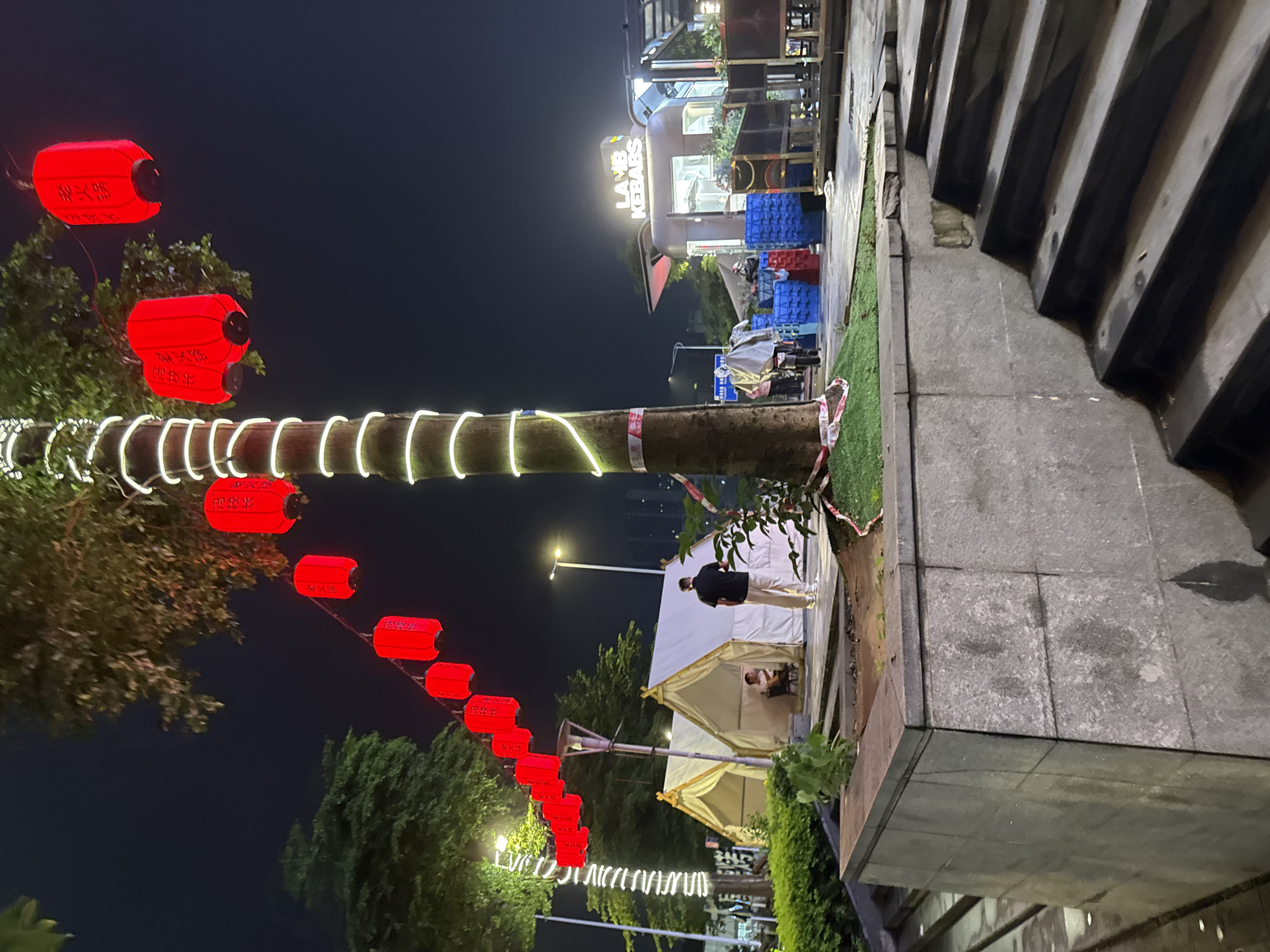 The image depicts an outdoor nighttime scene in Haizhu District, China, likely a market or entertainment area. Red lanterns hang in strings from trees, illuminating the area with a warm glow. Several white tents are set up, suggesting temporary stalls or seating areas. In the foreground, a person in a dark shirt and light-colored pants stands facing away from the camera, looking towards the tents. Further in the background, a food stall with a sign reading "LAMB KEBABS" is visible. The overall atmosphere is lively and festive, characteristic of a night market.
